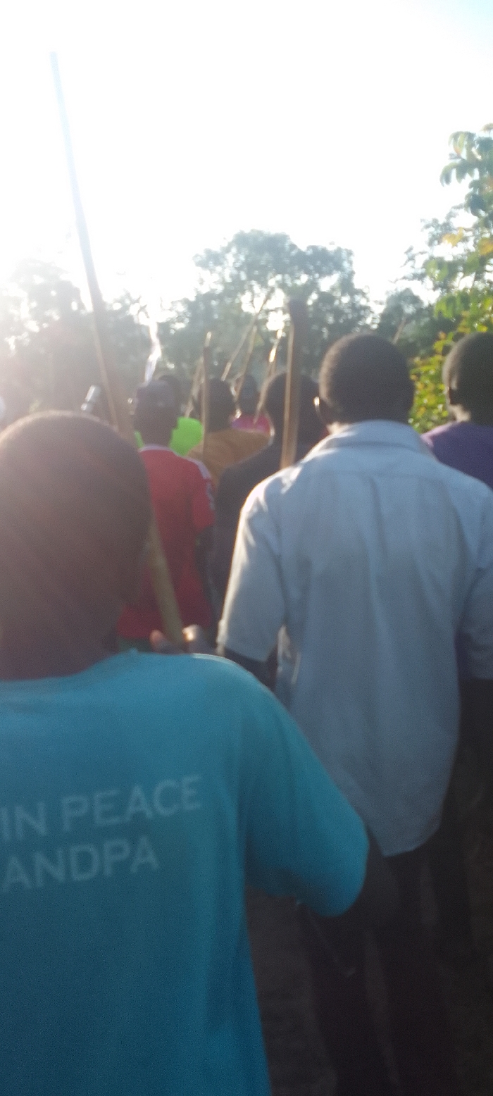 The image shows a group of people in a procession, with the individual in the foreground wearing a teal blue shirt that reads "IN PEACE ANDPA". The individuals in the background are carrying sticks and appear to be walking in a line. The scene is in a rural setting with trees in the background. The sky is bright and there is a lot of sunlight. The location is near Isukha ICHINA, Kenya, based on the provided coordinates. The individuals in the background are mostly obscured and out of focus. The overall feel of the image is peaceful and somber.