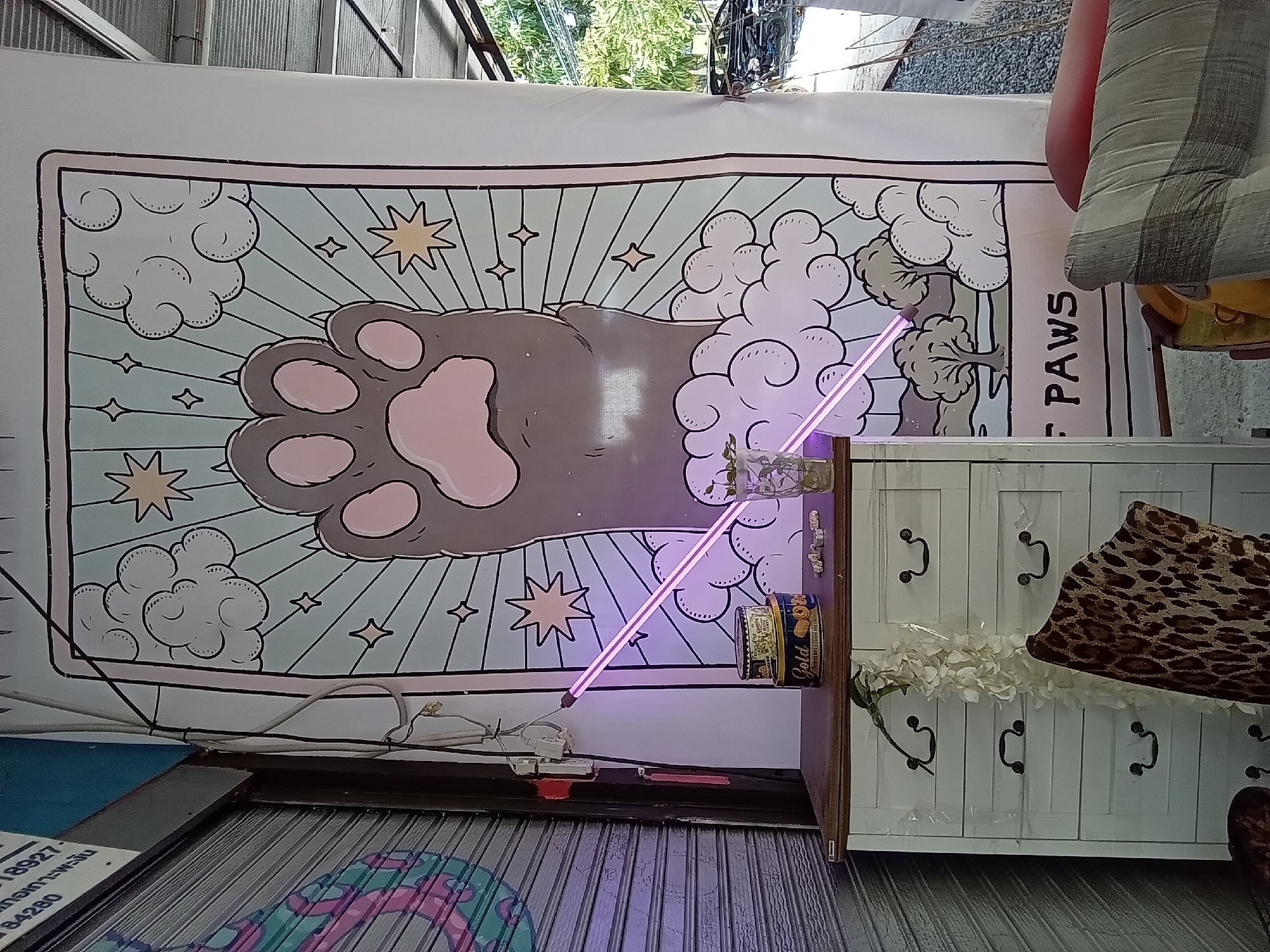A large banner depicting a cat's paw superimposed over a starry, cloudy sky with a sunburst pattern is displayed outdoors. Below the paw, the text "F PAWS" is visible. To the left of the banner, a white chest of drawers with dark drawer pulls is positioned. A white floral garland is draped over the chest, and a leopard-print chair is partially visible behind it. On top of the chest, two tin cans and a glass containing a plant are present. A pink neon light tube is diagonally positioned in front of the banner and chest. In the background, a rolled-up metal security gate with a colorful design is on the left, and a portion of a building with a corrugated roof and some trees are visible on the right. The scene appears to be in an outdoor commercial or retail area in ตำบลเกาะพะงัน, Thailand, given the context of the location provided, with evidence of a street or pathway in the background.