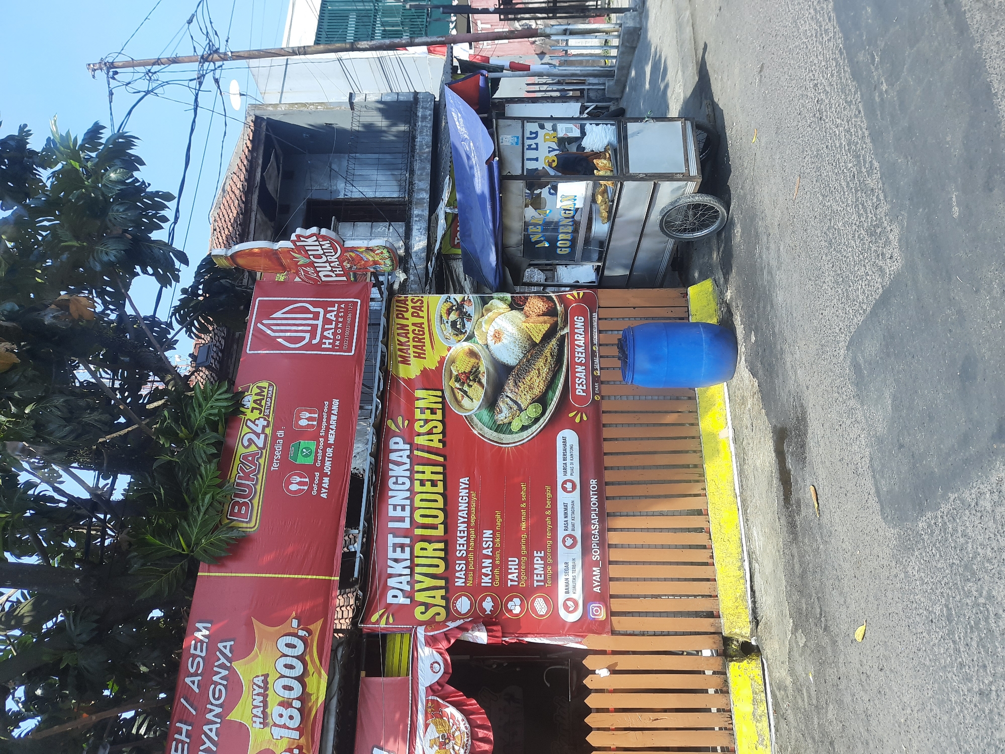 The image shows an outdoor scene in Bandung City, Indonesia, featuring a food stall and banners advertising food. In the foreground, a wooden fence with a yellow base runs horizontally. To the left of the fence, a large blue barrel stands on the asphalt. A food cart with a blue awning and metal structure is positioned to the right of the barrel. The cart displays fried food items. Behind the cart and fence, banners are hung. The topmost banner, predominantly red, has text including "BUKA 24 JAM" (Open 24 Hours) and mentions delivery services like GoFood and GrabFood, along with a location "AYAM JONTOR, MEKARWANGI". Another red banner below it advertises "PAKET LENGKAP SAYUR LODEH / ASEM" (Complete Package Sayur Lodeh / Asem), listing menu items such as "NASI SEKENYANGNYA" (filling rice), "IKAN ASIN" (salted fish), "TAHU" (tofu), and "TEMPE". An image of the food set is visible on this banner. A tree with dense foliage is prominent in the upper part of the image, casting shadows. The background shows buildings and a utility pole with multiple wires. The sky is clear and blue.