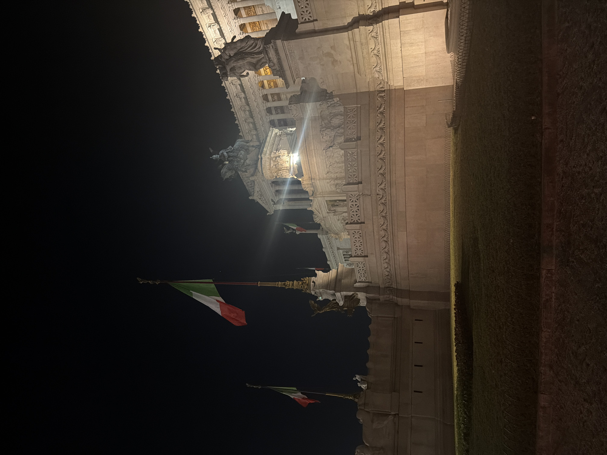 An exterior, eye-level, medium shot captures a nighttime view of a large, ornate stone building illuminated by artificial lights. Two Italian flags are visible, one larger and closer to the foreground on the right, and a smaller one further to the left. The flags are on tall poles and appear to be gently waving. The building itself has classical architecture with columns and intricate carvings. Statues are visible atop the structure, adding to its grandeur. A grassy area with some low-lying bushes is in the foreground, bordered by a low fence. The overall lighting creates dramatic shadows and highlights, emphasizing the texture and form of the architecture. The sky is dark, indicating nighttime.