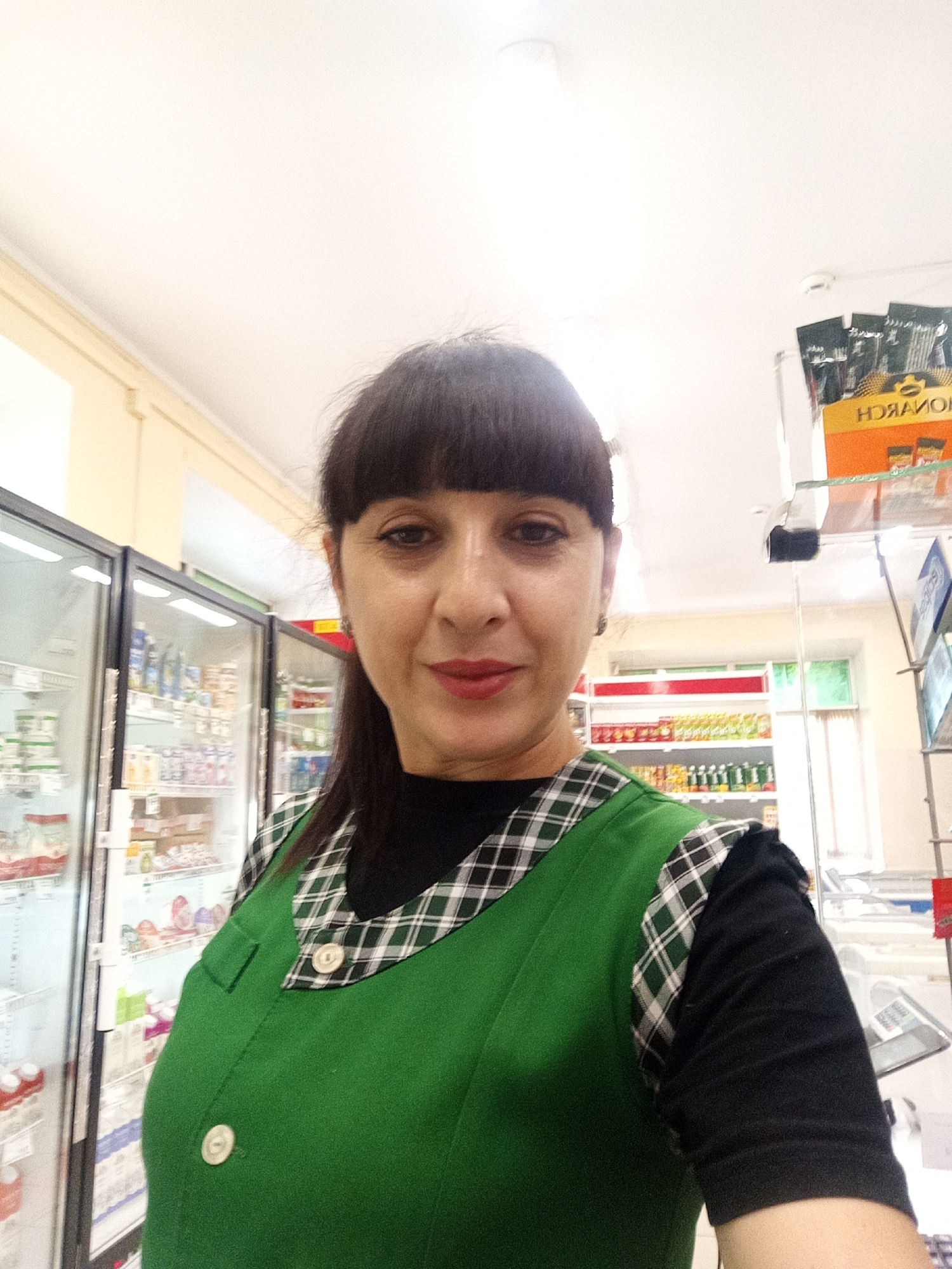 A woman with dark hair and bangs, wearing a green vest over a black shirt, stands in front of refrigerated display cases filled with products, likely in a grocery store. She is looking at the camera. The environment appears to be a commercial establishment with shelving and products visible in the background. The location is specified as Belogorsk, Russia.