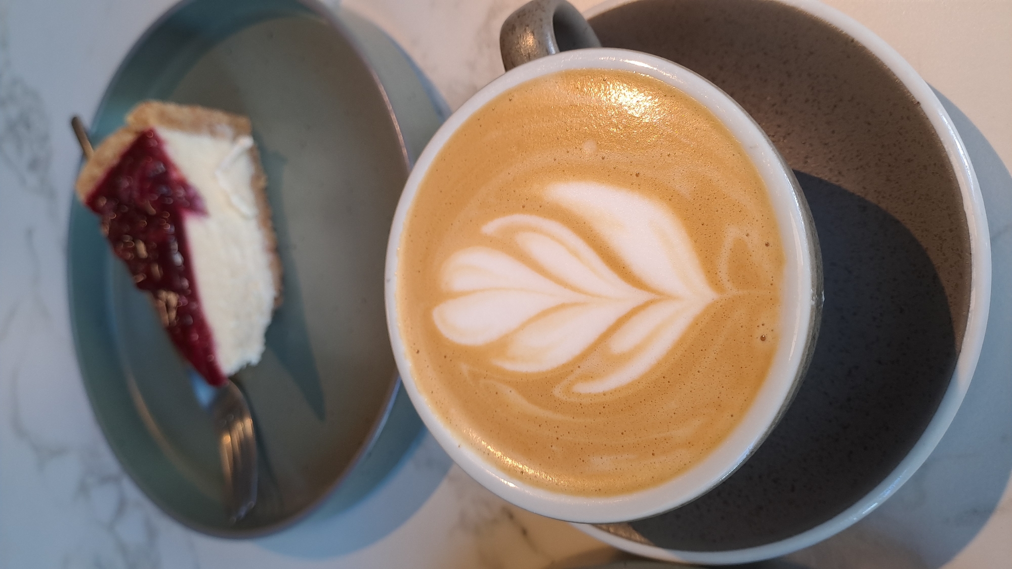 The image shows a close-up of a latte with latte art, placed on a dark gray saucer, which is itself on a lighter gray saucer. The latte art is a symmetrical tulip design. In the background, out of focus, is a slice of cheesecake topped with red berries, resting on a teal-colored plate. A fork is visible on the plate, partially obscured by the cheesecake. The surface beneath the plates appears to be a marble-patterned countertop. There are no people visible in the image. The image composition suggests a cafe or restaurant setting in Kolomyia, Ukraine, focusing on a dessert and coffee presentation.