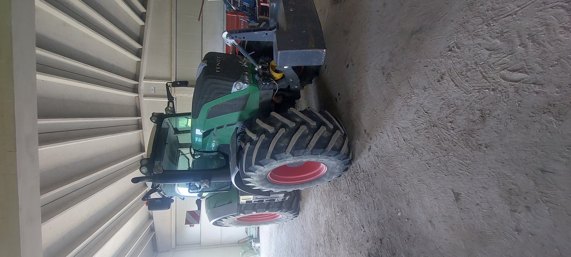 A green Fendt 828 Vario tractor is parked inside a covered structure. The tractor has large, rugged tires with red rims. It is oriented with its front facing slightly to the right of the camera, and its left side is more visible. A black counterweight is attached to the rear of the tractor. The interior of the structure appears to be a barn or workshop, with a concrete floor and a corrugated roof supported by beams. In the background, other farm equipment and supplies are visible, including what appears to be red machinery and possibly a pallet. The lighting suggests it is daytime. The location is identified as Pontestura, Italy.