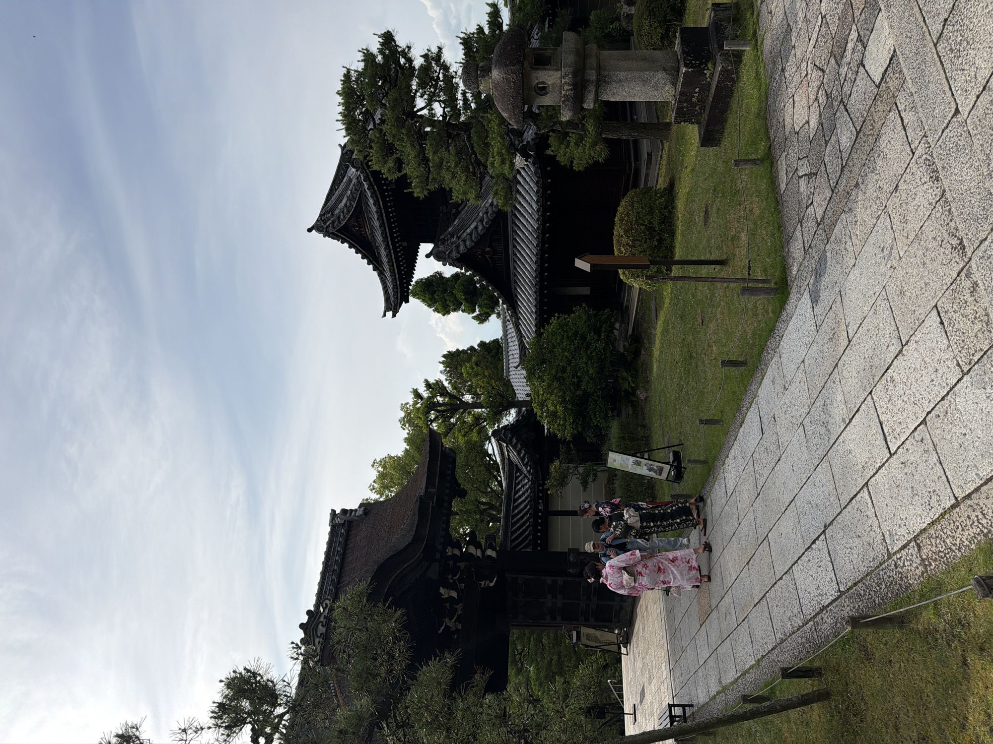 This image depicts an outdoor scene at Honen-in Temple in Kyoto, Japan, under a partly cloudy sky. A stone paved path extends from the foreground towards a large, dark wooden gate with a thick, rounded thatched roof.
On the path, four individuals are present. One female, wearing a pink kimono with a floral pattern and black sandals, walks away from the camera. To her left, two individuals stand facing each other: one female in a dark, patterned kimono, and a male in a light-colored cap, light jacket, and dark pants. A fourth female, also in a dark patterned kimono, is partially visible behind this pair.
To the right of the path, a manicured garden area features green grass, various shrubs, and pine trees. A weathered, hexagonal stone lantern stands prominently in the mid-ground. Further into the garden, a dark wooden, two-story traditional Japanese building with multiple tiled roof tiers is visible, flanked by trees. Another single-story dark wooden building with a tiled roof is partially obscured behind the two-story structure. Two black easel signs are positioned in the grass to the right of the path. On the far left, a dark wooden bench is partially visible. The stone path is bordered by low rope fencing supported by wooden posts. The sky above is light blue with scattered white clouds.