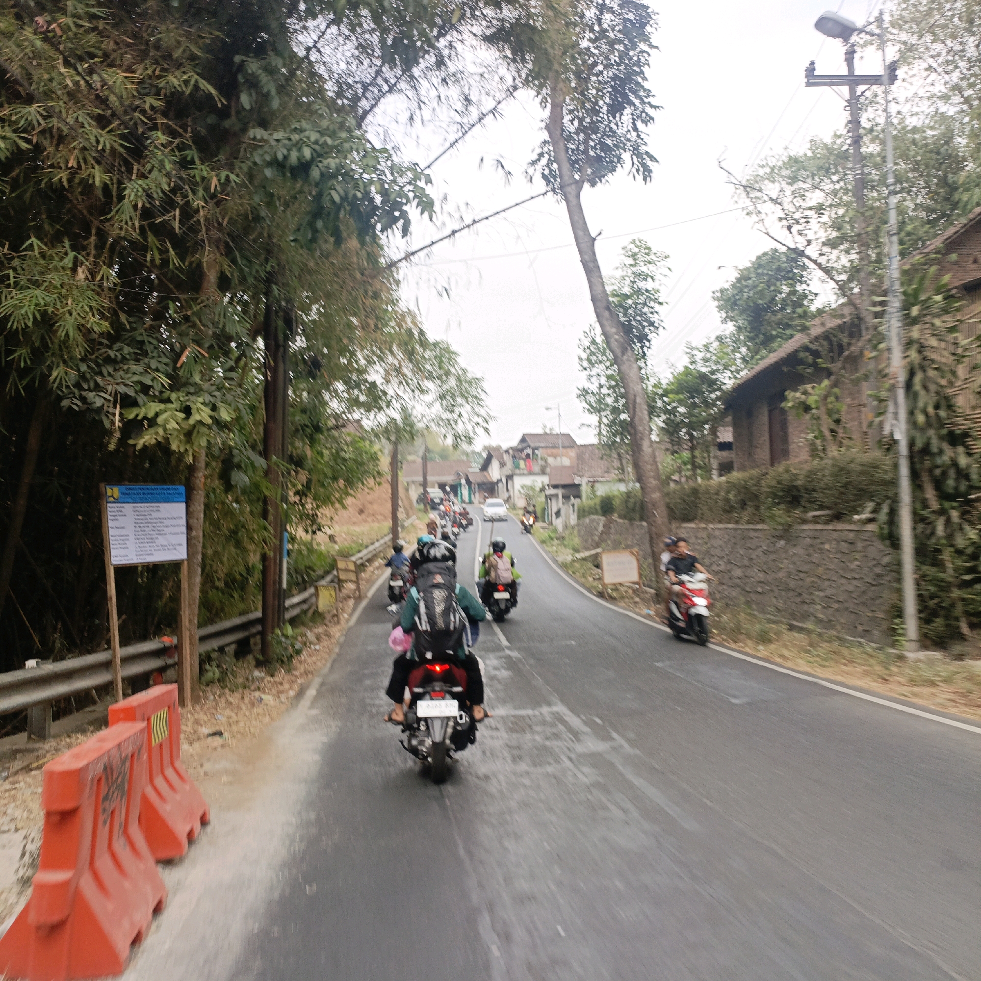The image depicts a street scene in Salatiga, Indonesia, likely during daylight hours, possibly in the late morning or early afternoon given the overcast sky. The perspective is from a moving vehicle, creating a sense of motion blur, particularly in the foreground and along the sides of the road.
The road is paved and appears to be a two-lane road winding through a semi-rural or suburban area. On the left side of the road, there are bright orange traffic barriers. Behind the barriers, a metal guardrail runs along the edge of a slope covered in vegetation, including bamboo trees and other foliage. A signpost with a blue and white informational sign is also visible near the barriers.
In the foreground, the rear of a motorcycle is captured, indicating that the photograph was taken from another motorcycle or vehicle following behind. The rider of this lead motorcycle is wearing a dark jacket and a backpack.
Further down the road, a procession of motorcycles is moving away from the viewer. Riders are visible on several of these motorcycles, some with passengers. The motorcycles are diverse in color and style, and some riders are wearing helmets. The road curves to the right, and in the background, several houses with tiled roofs are visible nestled amongst trees and vegetation. A white car is also seen in the distance, moving in the same direction as the motorcycles.
On the right side of the road, a stone wall runs along the edge of a property. A building with a brown, sloping roof and stone walls is situated behind this wall, partially obscured by trees and other greenery. A utility pole with a streetlight and power lines stands tall on the right side of the road. The overall impression is a typical Indonesian road with local traffic and residential areas.