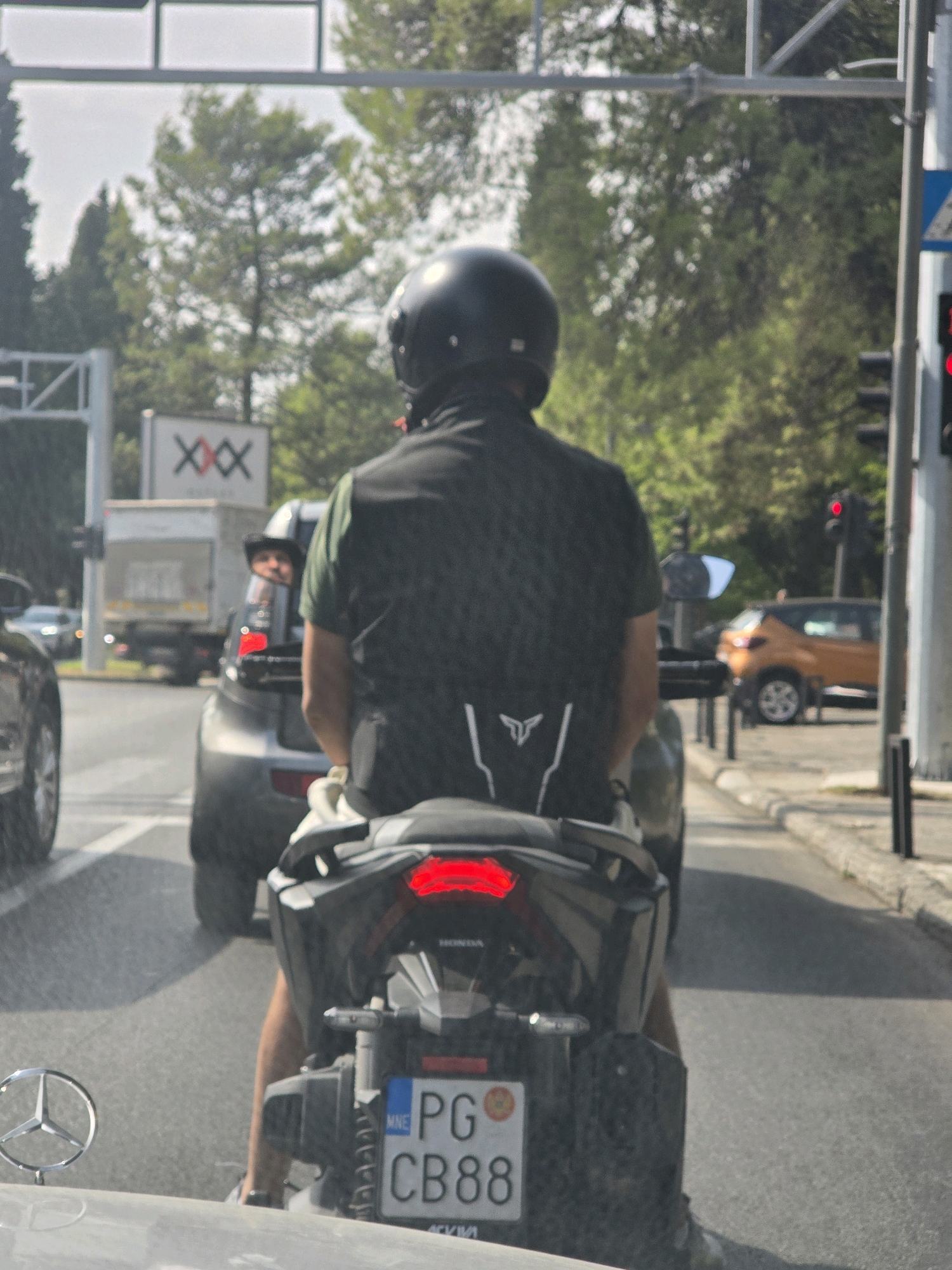 A rear-view, eye-level, medium shot captures a person riding a scooter in traffic on a sunny day. The rider, wearing a black helmet and a dark green vest over a black shirt, is facing away from the camera. Their legs are visible, clad in dark trousers and white-laced sneakers. The scooter's license plate is prominently displayed, showing "PG CB88 MNE".
The scooter is positioned behind a gray car, and to its left, another car's wheel is visible. In the background, traffic signals are visible, with a red light illuminated on the right. An orange car is parked on the right side of the road. Trees line the background, suggesting an urban or suburban environment. In the far background, a large truck with "XXX" marked on a sign is visible. The scene appears to be an intersection or a busy street in Podgorica, Montenegro, indicated by the "PG" on the license plate, which is a prefix for Podgorica. The lighting suggests it is daytime, with bright sunlight casting shadows.