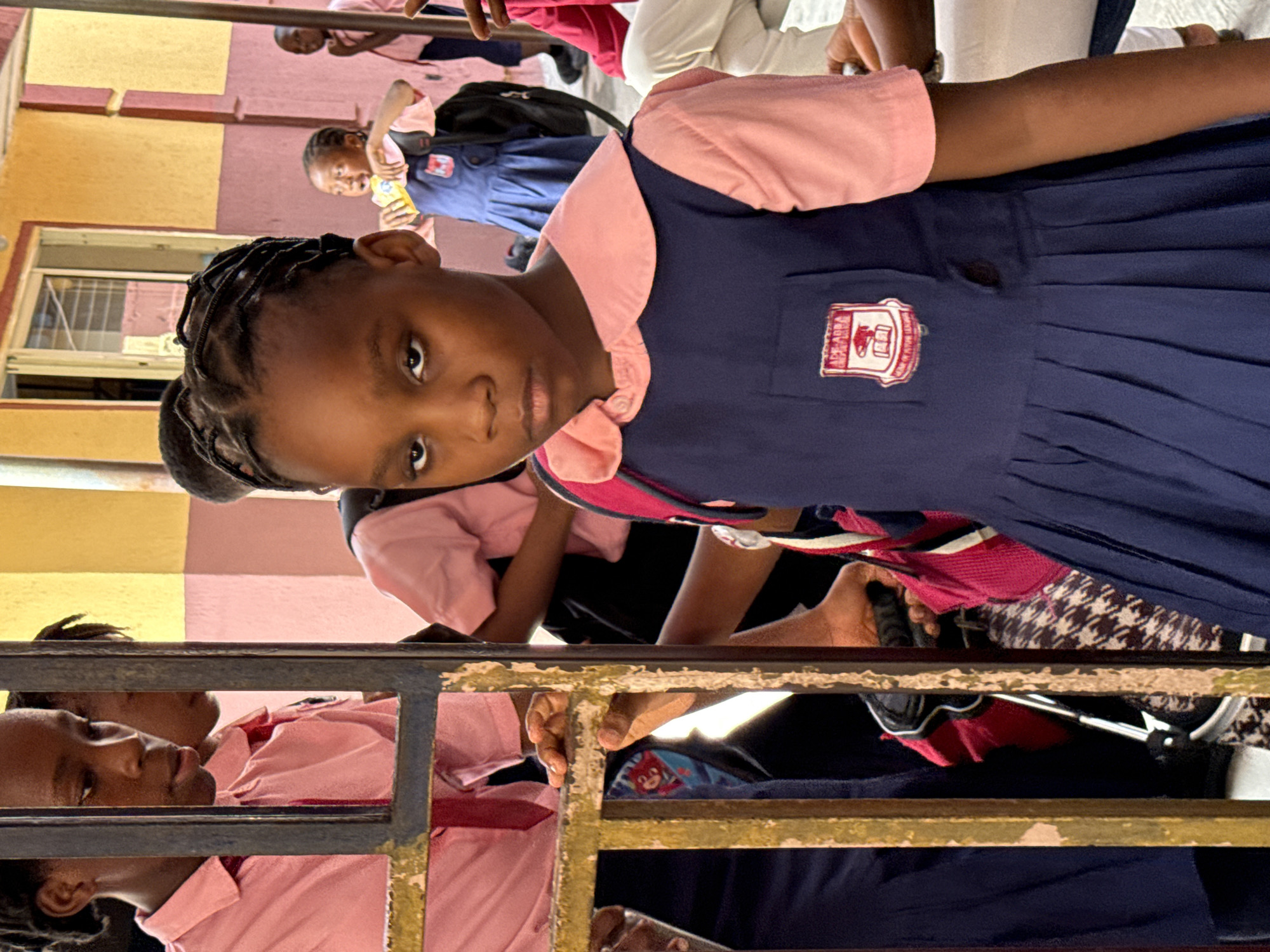 The image shows a group of schoolchildren in Ado, Nigeria, primarily girls, wearing a navy blue and pink school uniform. The main subject is a girl in the foreground, looking directly at the camera with a serious expression. She is wearing a school uniform with a badge on the chest. In the background, other children can be seen, some also wearing the same uniform, and others in different attire. Some of the children are behind a metal railing. The environment includes a building with yellow and pink colored walls, suggesting an institutional setting, likely the school premises.