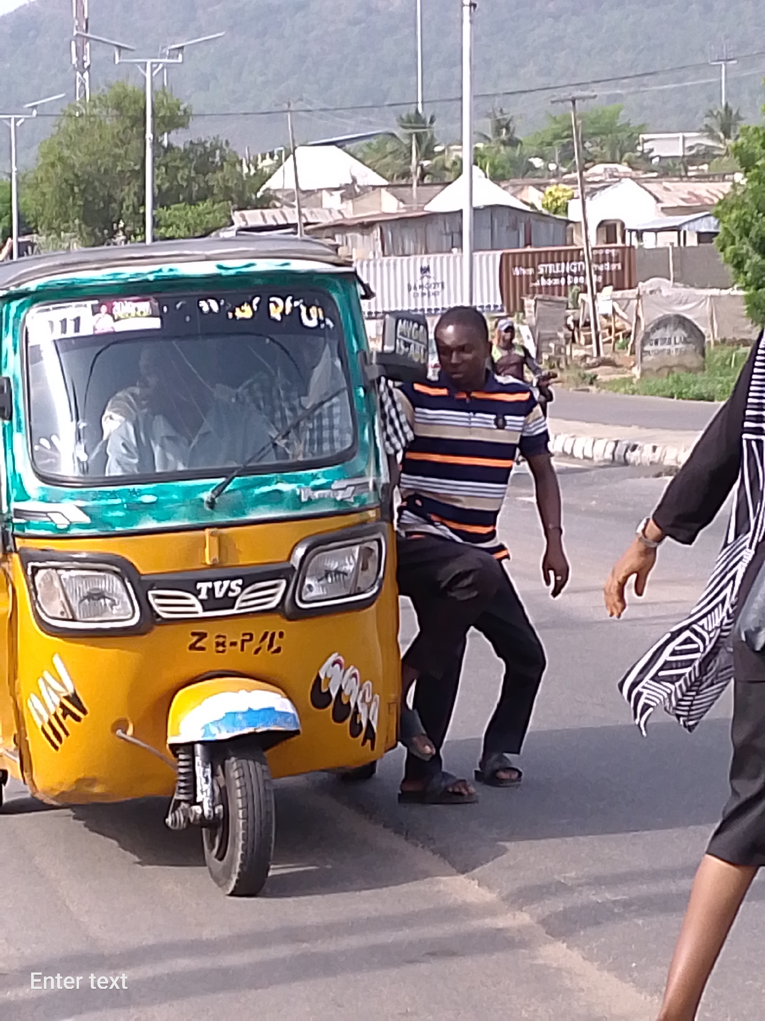 In Adankolo, Nigeria, a yellow and green "Keke Napep" (three-wheeled auto rickshaw) is parked on an asphalt road. A man wearing a striped shirt and black pants stands beside the vehicle, resting his hand on it. The auto rickshaw is partially adorned with painted decorations, including the words "ONE WAY" on the left side and what appears to be a repeated word on the right. A person is visible inside the vehicle. Another person is partially visible on the right edge of the image, wearing a dark outfit with a patterned scarf. The background shows buildings, shipping containers, trees, and mountains under a daytime sky.