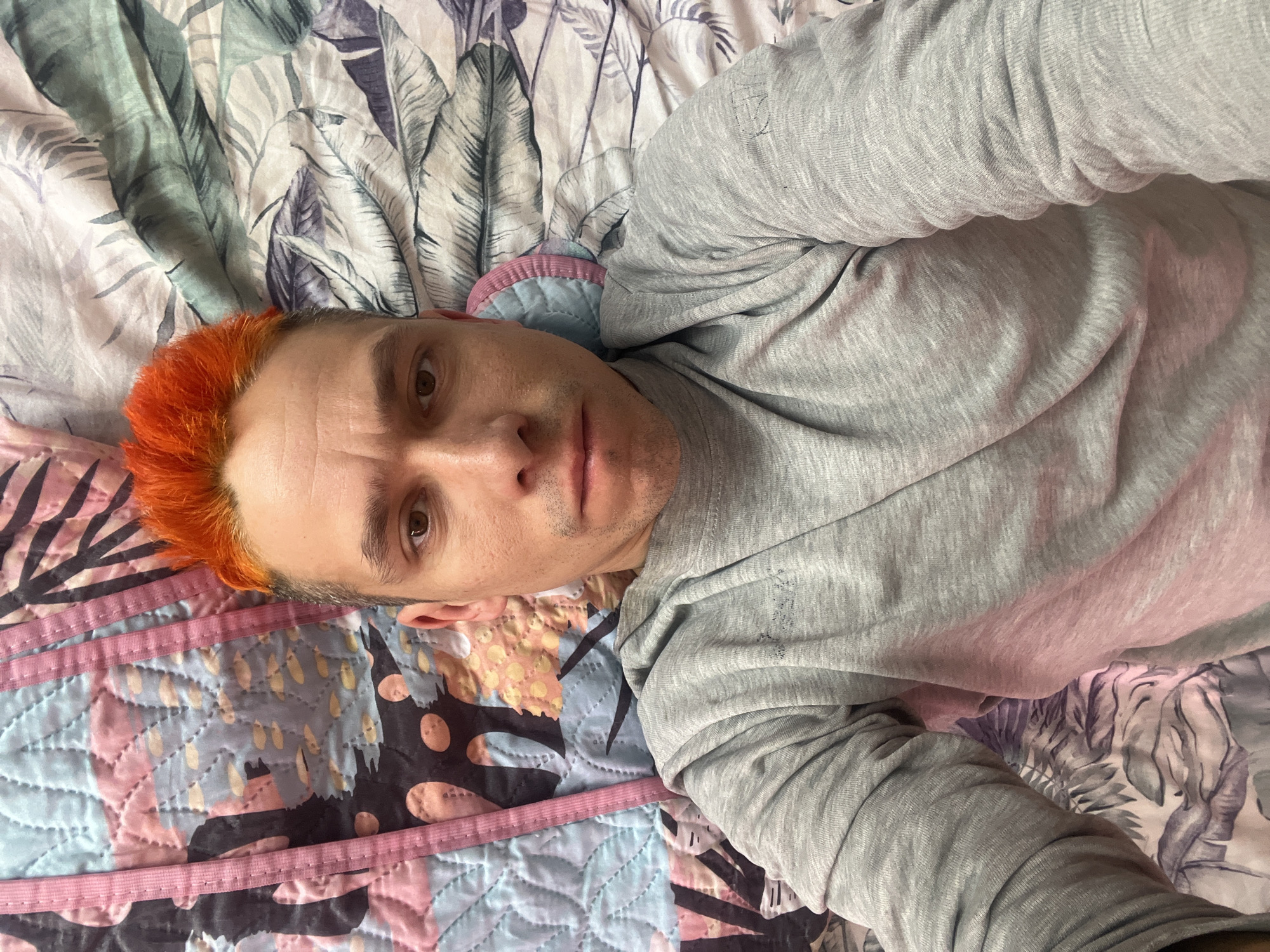 A medium shot captures a man lying on his back on a bed. The man has fair skin, brown eyes, and short orange hair. He is wearing a gray long-sleeved shirt. The bed has a colorful quilt with pink trim and a light-colored floral pattern on the top half. The location is Unknown, Unknown.