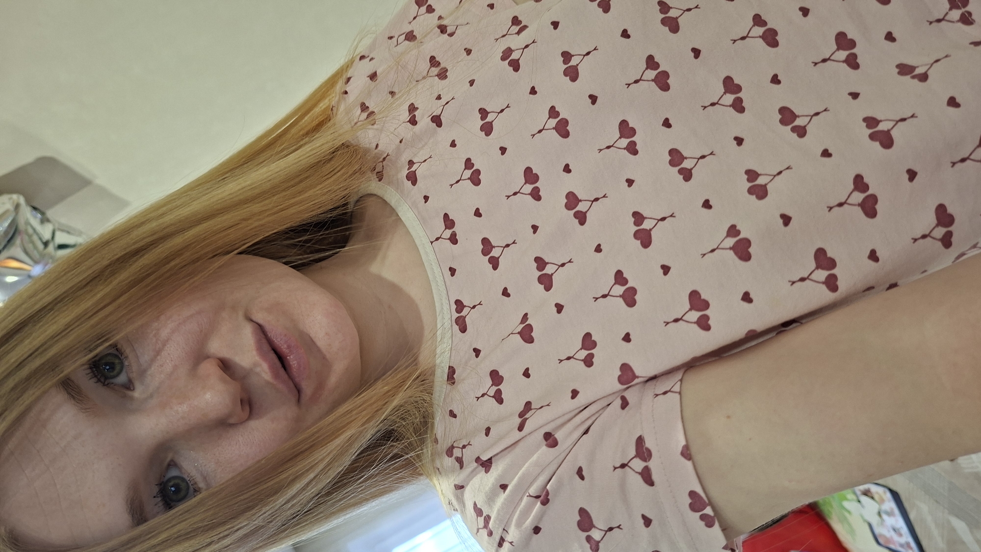 The image is a selfie of a light-skinned woman with blonde hair and blue eyes. She's wearing a light pink top with a pattern of small, red heart-shaped balloons. The location is possibly in a domestic setting, and may be in the city of Chaikovsky, Russia. There are no discernible specific actions or interactions evident.