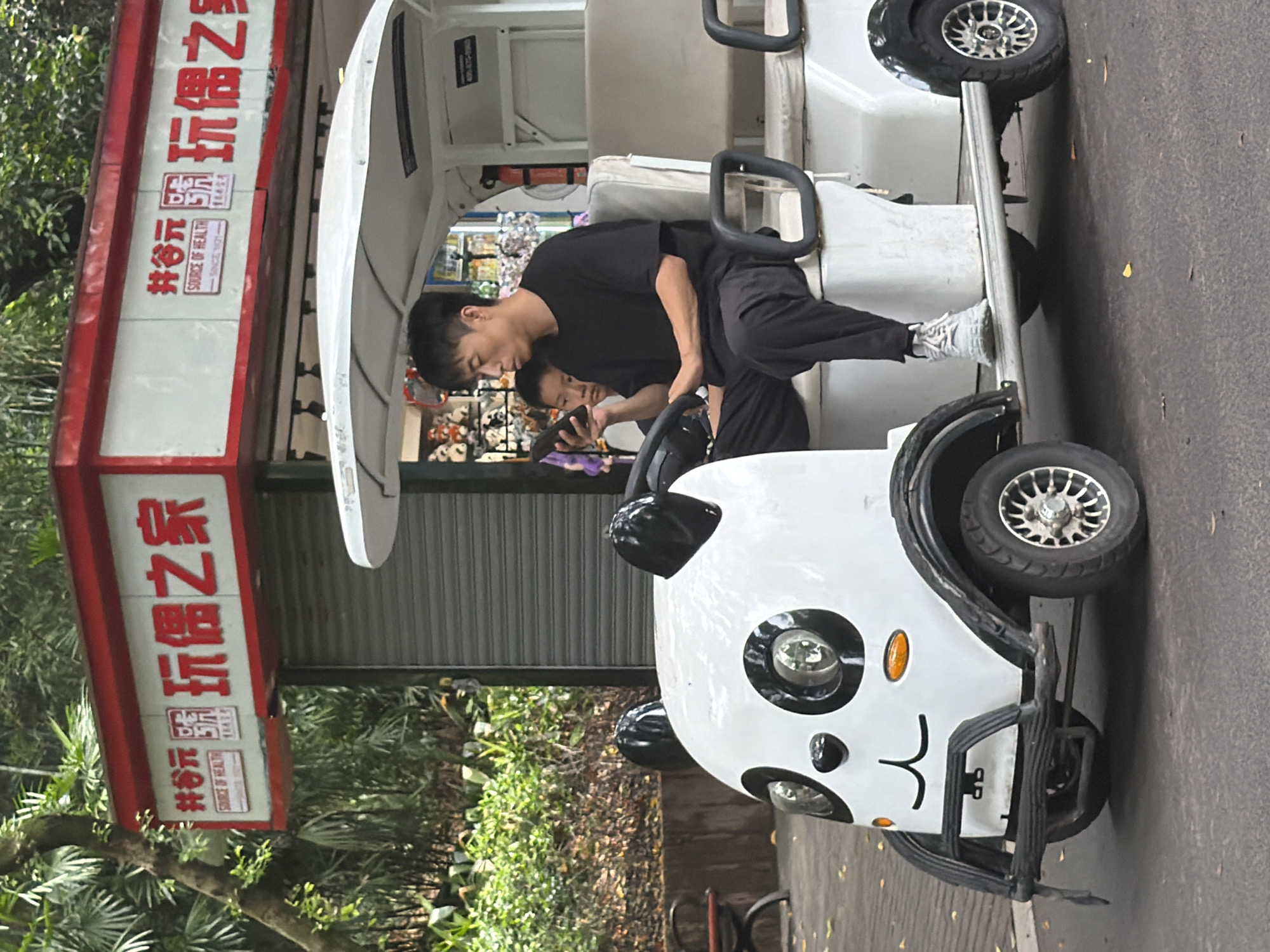 In Jiulongpo District, China, a white electric cart designed to resemble a panda is parked on an asphalt surface. The cart has large black panda ears on its roof and a black and white face with round headlights for eyes. A man, wearing a black t-shirt and black pants, is seated in the driver's seat of the cart, holding a black smartphone. Behind him, a young boy sits in the passenger seat, looking forward. The cart is positioned in front of a building with a red awning and a sign displaying Chinese characters and the English text "SOURCE OF HEALTH." To the left of the cart, lush green foliage and trees are visible, suggesting an outdoor, possibly park-like setting.