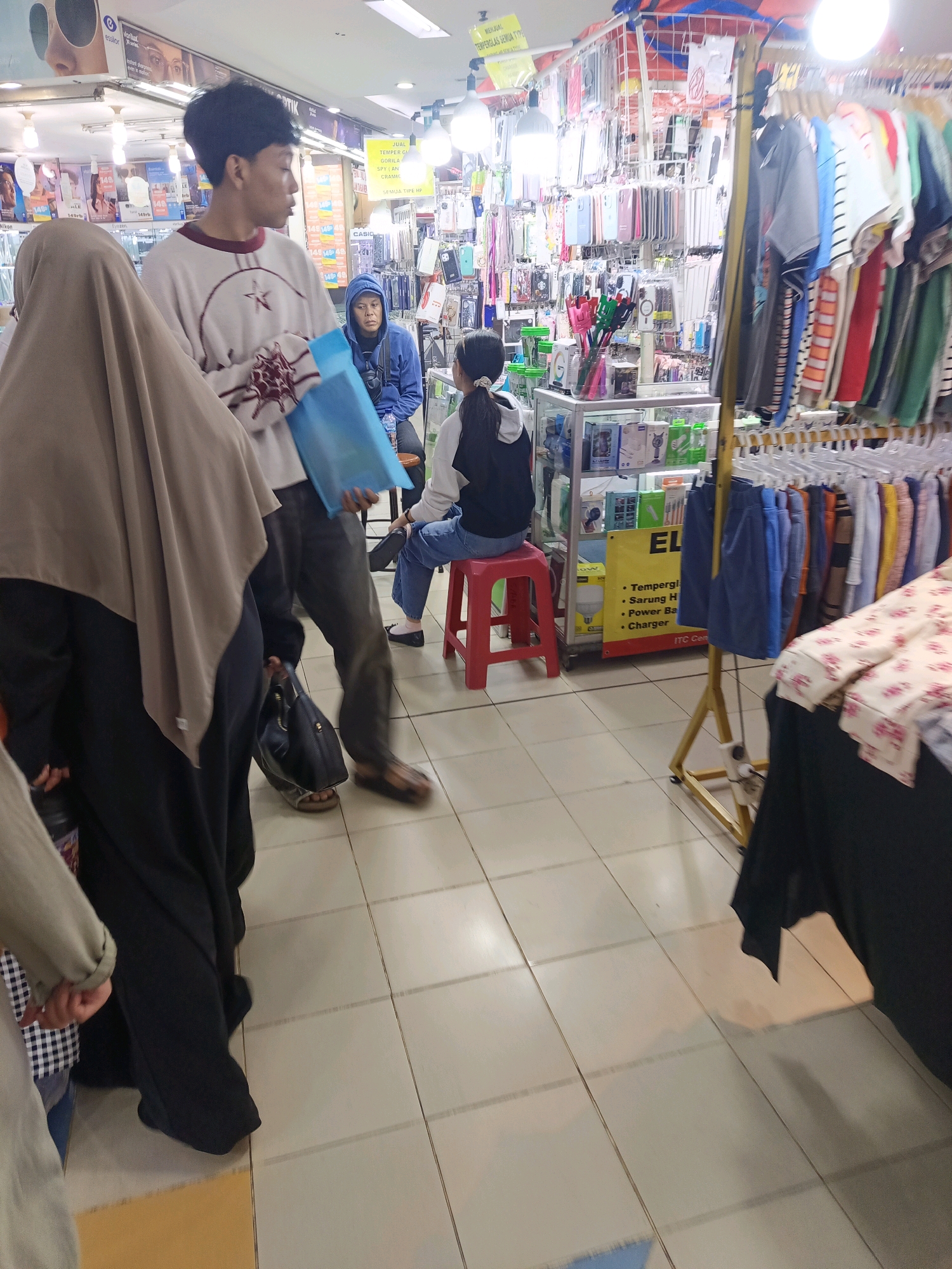 The image captures an indoor scene, likely a market stall or a shop in Central Jakarta, Indonesia, given the signage that appears to be in Bahasa Indonesia. The foreground is dominated by the lower bodies of several individuals. A person on the left is wearing a black abaya and a beige hijab. Next to them, a person in a white sweatshirt with a maroon collar and a star-like design is holding a blue plastic bag. Another person, wearing a blue hoodie, is seated on a red stool to the right. Further to the right, a person in a black hoodie and blue jeans is also seated on the red stool. The background reveals racks of clothing, shelves stocked with mobile phone accessories like cases and chargers, and various signs and advertisements. The floor is tiled with light-colored square tiles. The overall impression is a busy commercial environment.