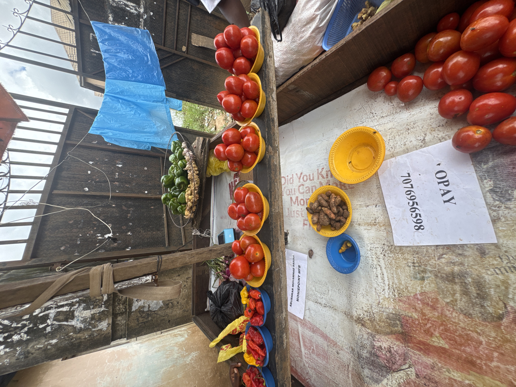 A street vendor in Ihiagwa, Nigeria displays produce for sale. The stall features several bowls of red tomatoes, as well as bowls containing red and yellow chili peppers. In the background, a basket holds green bell peppers, and ginger roots are also visible. A blue plastic sheet is strung up behind the stall, and a wooden fence encloses the area. A price list or sign with the text "ABUBAKAR MUHAMMAD SARINA MONIEPOINT MFB" and a phone number "8656598707" is visible. The ground is covered with a patterned cloth. The scene appears to be outdoors, with a cloudy sky overhead.