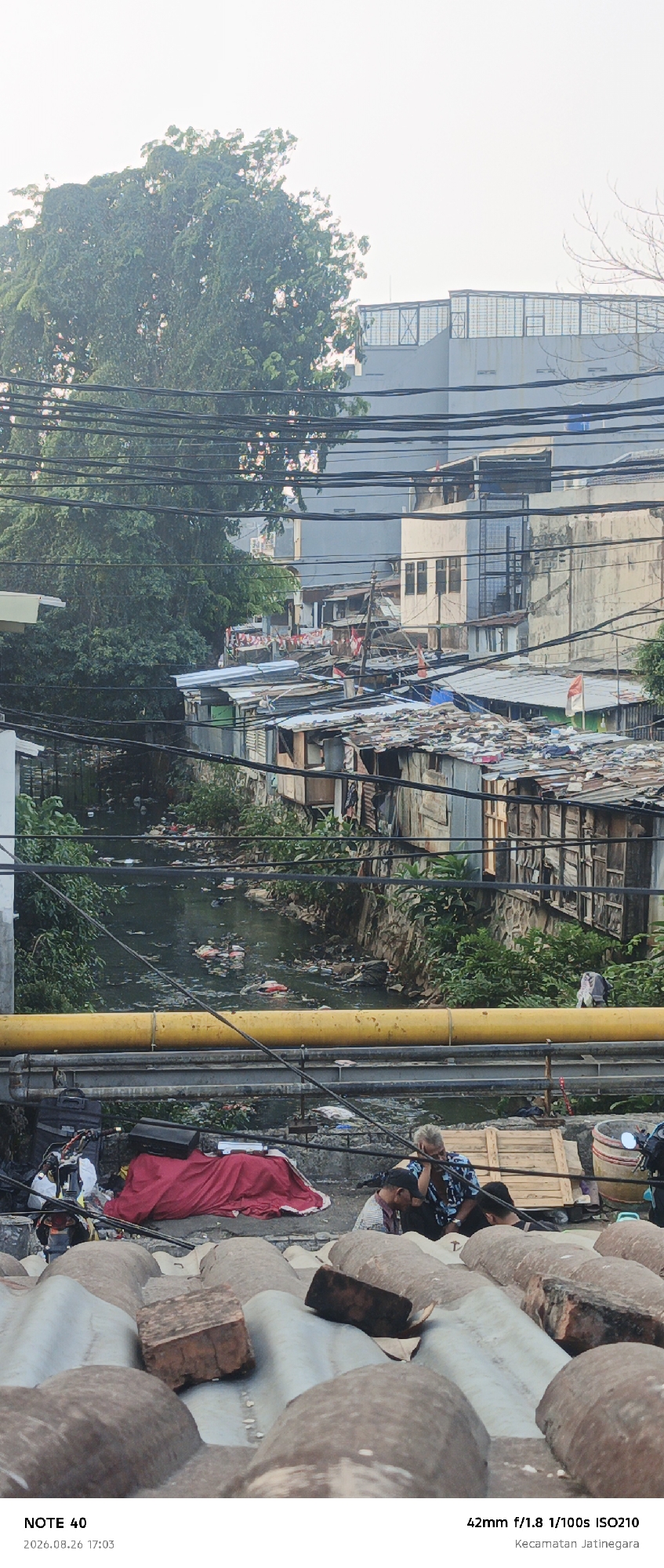 The image depicts a view from an elevated position, looking down onto a canal filled with debris and a densely populated area with makeshift structures. In the foreground, a corrugated roof is visible, with two bricks on top of it. Below the roof, several people are gathered. A man with blond hair is seated, looking down, with two other individuals near him, one appearing to be a child. A red cloth is spread out on the ground, and various items are scattered around.
The canal, which runs through the scene, is murky and filled with trash. The banks are lined with vegetation, but the water itself is heavily polluted. On the opposite side of the canal, a cluster of informal dwellings, constructed from corrugated metal and wood, are densely packed together. Some of these structures have flags hanging from them, indicating a national celebration. In the background, modern buildings rise above the informal settlement, creating a stark contrast between development and poverty.
The overall scene suggests an urban environment in East Jakarta, Indonesia, specifically in the Kecamatan Jatinegara area, characterized by both modern infrastructure and the presence of informal settlements along a polluted waterway. The image captures a moment in time with people gathered in the foreground, possibly in a residential area. The lighting suggests it is either early morning or late afternoon.