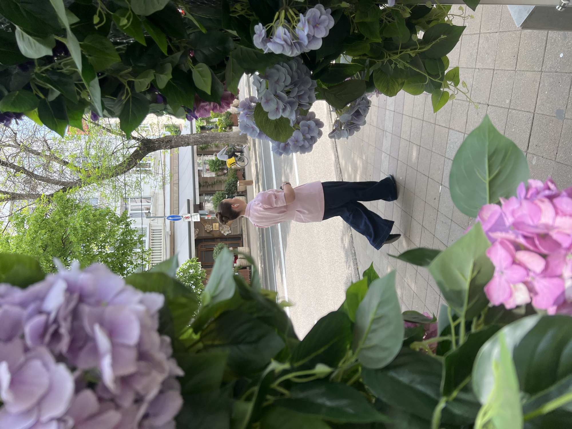 A young woman is standing on a sidewalk in Krasnodar, Russia, framed by flowering plants. She is wearing a pink shirt and dark blue pants. A tree and buildings are visible in the background. The sidewalk is made of light gray pavers. A cyclist is in the background.