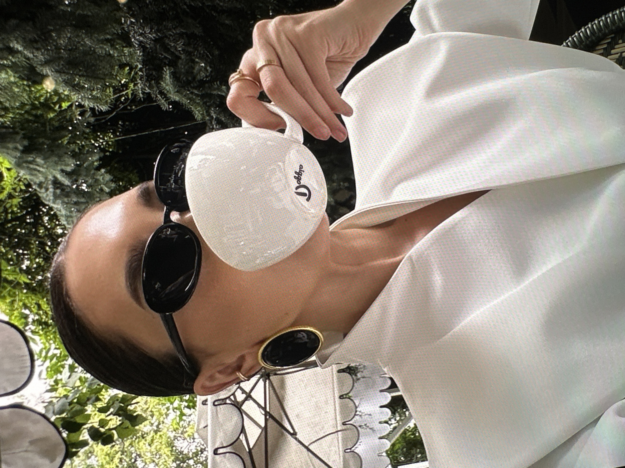 A woman with dark hair pulled back and wearing large black sunglasses and gold hoop earrings is holding a white coffee cup with the word "Doppio" printed on it. She is wearing a large, structured white garment. The background includes trees and what appears to be decorative metalwork. The location is Yerevan, Armenia.