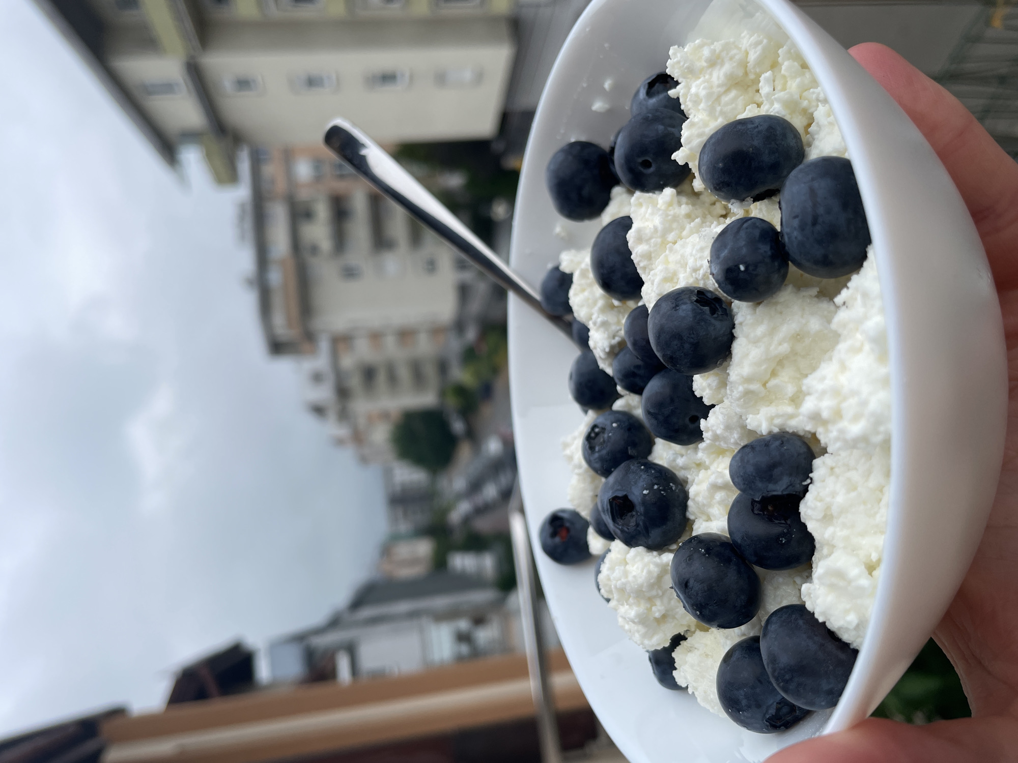 The primary subject of the image is a white bowl filled with cottage cheese or ricotta, generously topped with fresh, dark blue blueberries. A silver spoon is resting within the bowl, partially submerged in the food. A light-skinned hand is visible on the right, holding the bowl.
The food appears to be a healthy breakfast or snack. The cottage cheese is white and has a lumpy texture, while the blueberries are round and plump, some with small white specks on their surface.
In the background, out of focus, are several multi-story buildings, suggesting an urban or suburban setting. These buildings are light-colored with visible windows and architectural details. The sky above them is overcast and cloudy, indicating a non-sunny day. A blurred brown or beige surface, possibly a railing or a table, is visible in the lower part of the background, suggesting the photo was taken outdoors, perhaps on a balcony or terrace.
The image is oriented sideways, with the buildings appearing tilted or horizontal relative to the food. No text is visible in the image.