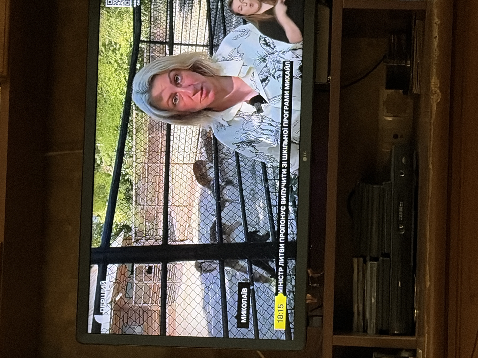 The image shows a television screen displaying a news broadcast. In the foreground, a woman with blonde hair, wearing a patterned shirt, is speaking. Behind her, a fenced enclosure with some animals, possibly deer, is visible. A smaller screen in the lower right corner shows a woman signing. The screen also displays text overlays, including the channel name "ПЕРШИЙ" at the top left, and what appears to be a location "МИКОЛАЇВ" and a news ticker at the bottom: "18:15 МІНІСТР ЛИТВИ ПРОПОНУЄ ВИЛУЧИТИ ЗІ ШКІЛЬНОЇ ПРОГРАМИ МИХАЙЛ". Below the television, a shelf holds several stacks of DVDs or CDs and a black electronic device, possibly a DVD player. The background of the room is dark and appears to be made of wood. The overall scene is an indoor setting, likely a living room, where someone is watching television.