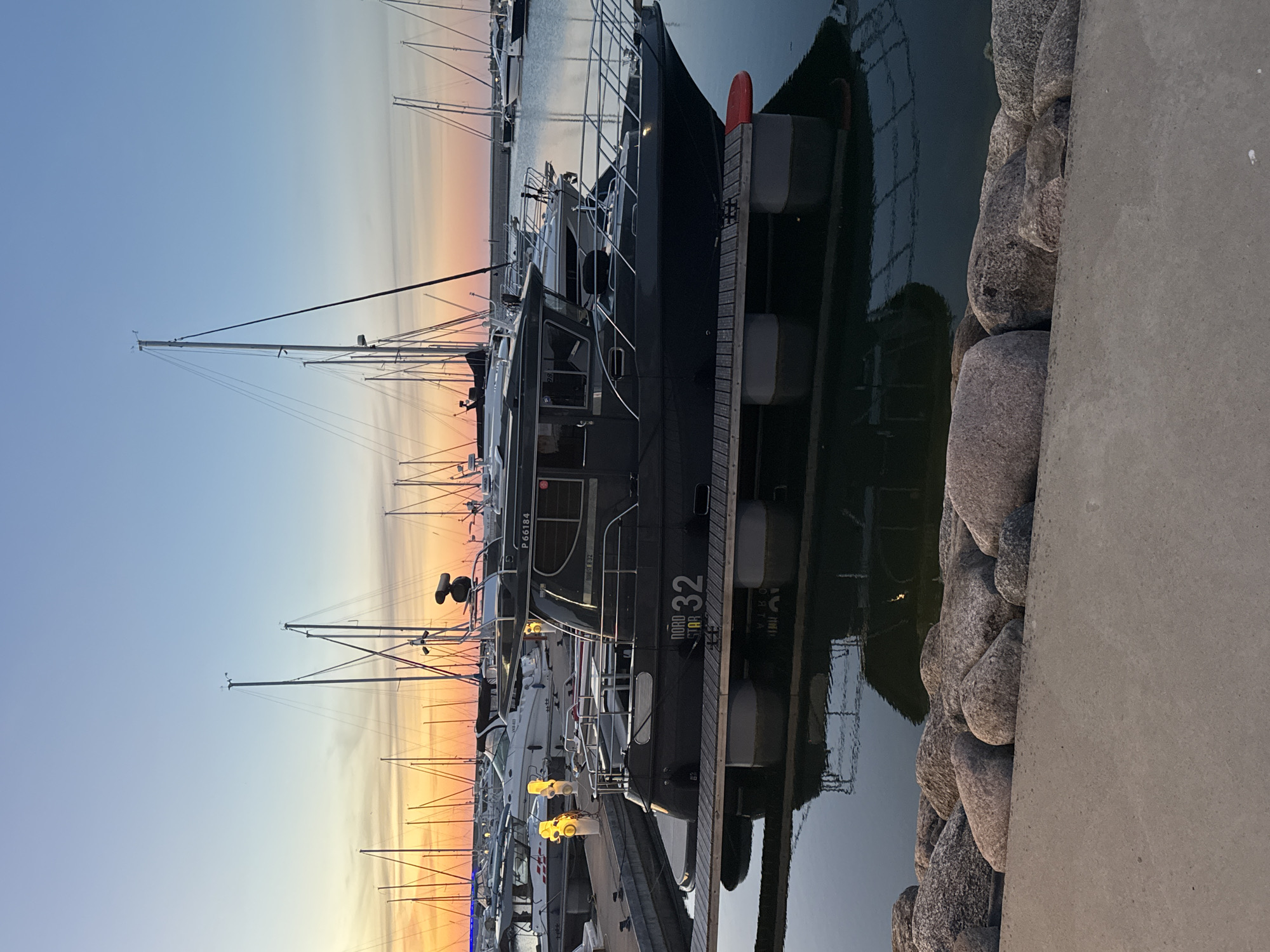 In Tallinn, Estonia, at a marina during sunset, a dark-hulled motorboat named "NORD STAR 32" is docked. The boat, with registration number P 66184, is positioned on a wooden platform that is supported by large cylindrical floats. The vessel features a cabin with windows and a metal railing around its deck. Behind this boat, several sailboats with tall masts are visible, their sails furled. The sky displays a gradient of colors from the setting sun, transitioning from orange and yellow near the horizon to a clear blue overhead. The water surrounding the boats is calm, reflecting the sky and the boats. In the foreground, a concrete walkway borders a section of large, rough-surfaced rocks. The scene is tranquil, with no people visible.