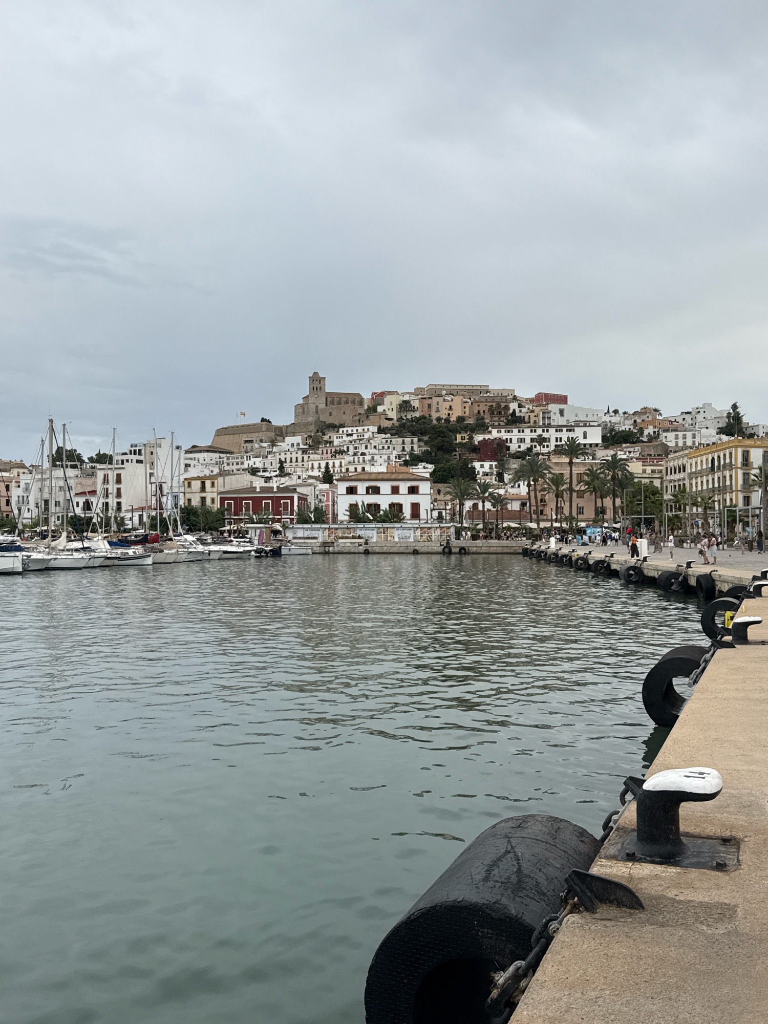 The image depicts a harbor scene in Ibiza, Spain, with the city's historic Dalt Vila fortress and its white-washed buildings visible in the background, perched on a hill. In the foreground, a concrete pier is lined with large, black fenders and mooring bollards. The water in the harbor is calm and reflects the overcast sky. Several sailboats are moored along the waterfront, their masts creating a vertical element against the backdrop of the town. People are walking along the pier in the middle ground, some engaging in conversation, while others are strolling casually. The architecture of the buildings in the town ranges from traditional white structures to some with colorful facades and terracotta roofs. Palm trees are interspersed among the buildings, adding a tropical feel. The overall atmosphere suggests a peaceful day in a Mediterranean port city.