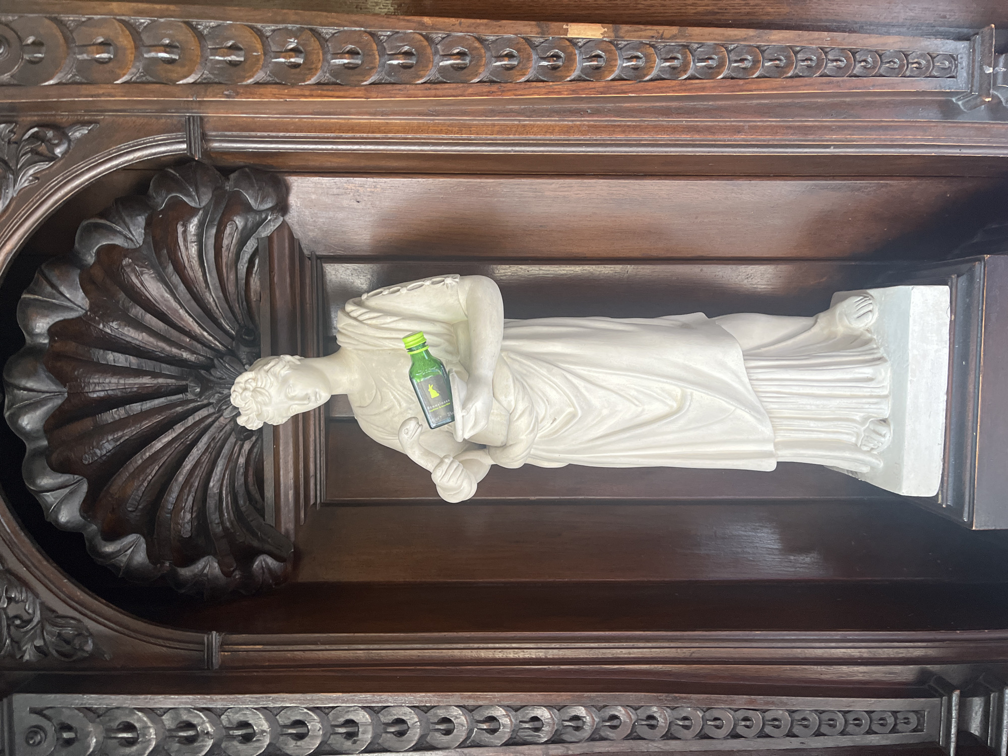 A white marble statue of a draped woman stands within a dark wood alcove. The statue is holding a small green bottle in its hands, which appear to be cupped. The woman has her hair in an updo. The alcove features ornate carvings, including a fan-shaped wooden element above the statue and decorative bands on the wooden panels to the sides. The background is dark wood.