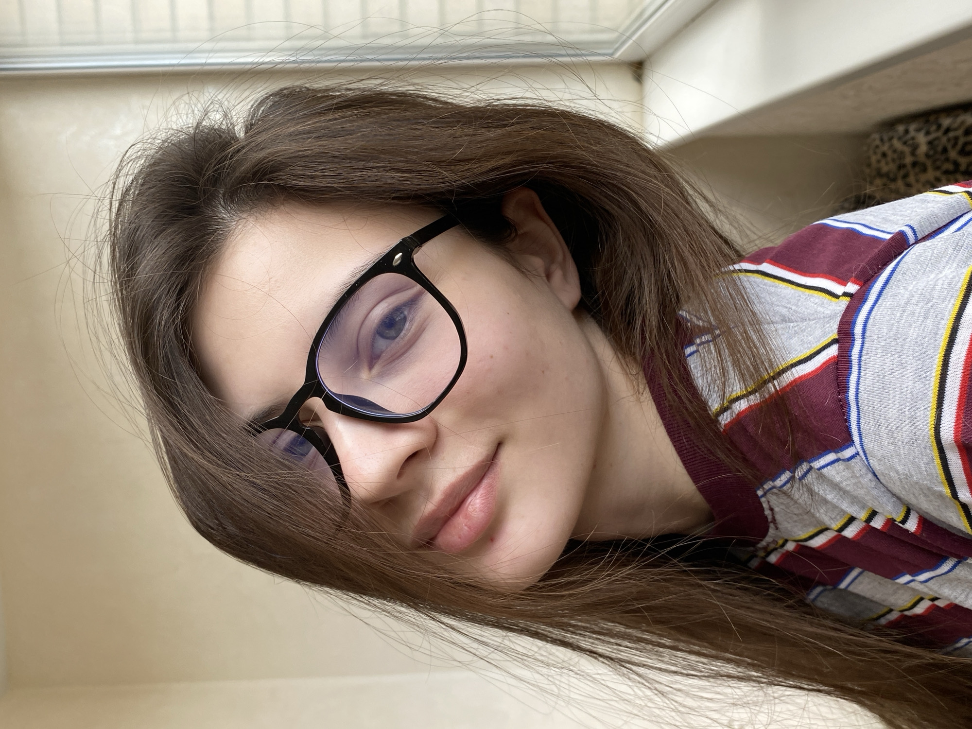 A close-up, rotated portrait features a young individual with long, straight dark brown hair and black-framed glasses. The subject's head is tilted, and they are looking directly at the camera with a gentle, soft smile, revealing light-colored eyes. They are wearing a striped top with a pattern of maroon, grey, blue, yellow, and red bands.
The lighting is soft and diffused, indicating natural daylight indoors. The background is largely a plain, light-colored surface, possibly a wall or ceiling, with a subtle hint of a leopard print pattern visible in the upper right portion, suggesting an indoor domestic setting. The image is framed as a low-angle shot, possibly a selfie.
The subject's expression appears calm and content. No visible text is present in the image. The provided location context for this media is Votkinsk, Russia.