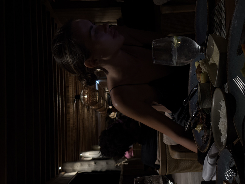 A woman with her hair up, wearing a black top, is seated at a dimly lit table. Plates of food and a glass of liquid with a lime wedge are present. A person with dark curly hair is visible in the background. The surroundings include textured walls and decorative hanging lights. The scene seems to be an intimate dinner in a restaurant with a warm ambiance.