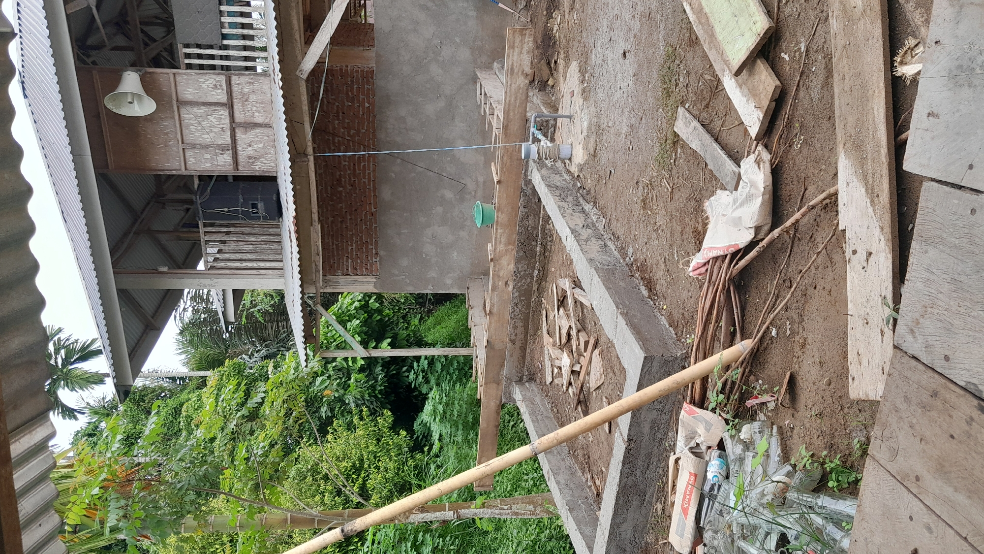 The image shows a construction site in Lhokjok, Indonesia. A newly constructed concrete foundation is visible. Building materials, including wooden planks, bamboo poles, and cement bags are scattered around. A partially constructed brick and concrete building stands adjacent to the construction area. Lush vegetation surrounds the construction site. A water pipe is visible, and a string with a green bucket hanging from it.
