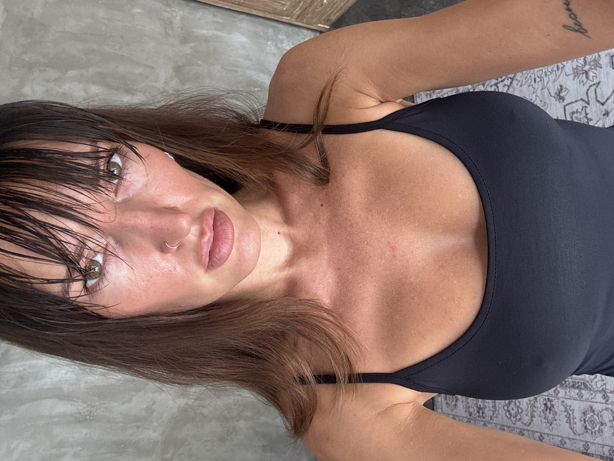 A woman with wet, dark brown hair is pictured indoors from a high angle. She has fair skin and is wearing a black top with thin straps. Her lips are tinted pink, and she has a silver nose ring in her left nostril. The background is a greyish surface, possibly a rug. The location may be North Kuta, Indonesia, as specified. A tattoo is visible on the woman's right arm.
