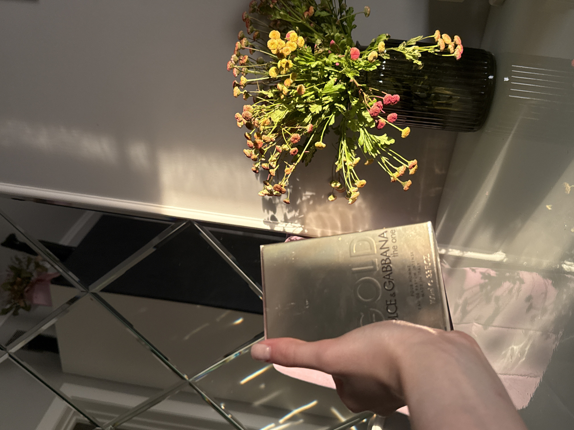 The image shows a close-up of a hand holding a box of Dolce & Gabbana "The One Gold" perfume. The box is a metallic gold color with black lettering. The hand has well-maintained fingernails. In the background, a bouquet of dried flowers in a dark, ribbed vase is visible. Sunlight is casting shadows on the wall behind the flowers, suggesting it is daytime. The scene appears to be indoors, possibly on a table with a reflective surface, as indicated by the diamond-patterned mirrored surface in the lower part of the image, which reflects the flowers and light. The overall impression is one of product presentation, possibly for advertising or a personal review, with a focus on the perfume box. The lighting creates a warm and somewhat dramatic effect. There is no clear indication of the specific location, but the setting appears to be a decorated interior.