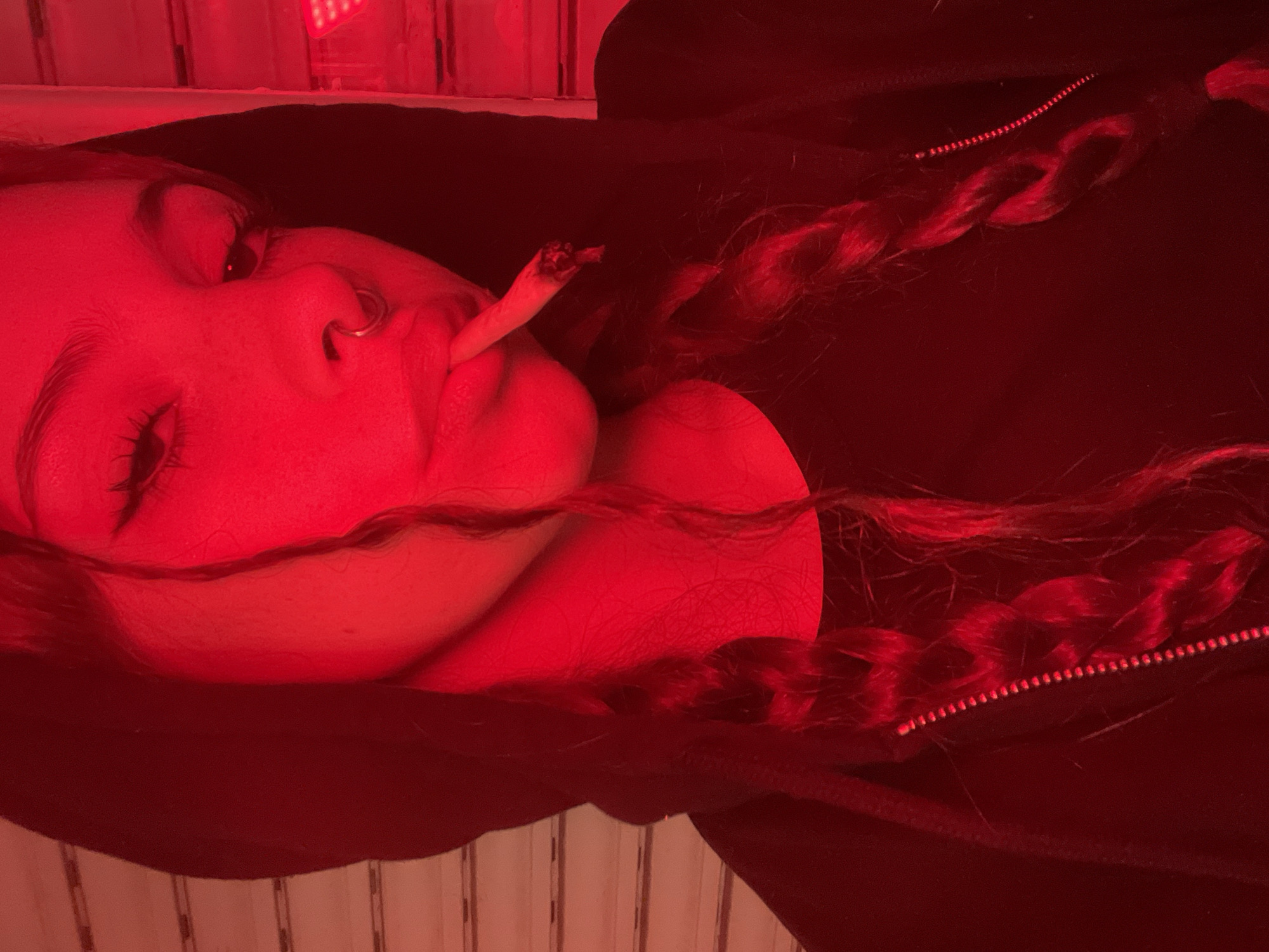 A young woman is lying down, facing slightly upwards to the left. She has braided hair, a nose ring, and is holding a lit cigarette between her lips. She appears to be wearing a black hooded jacket. The scene is lit by a strong red light, obscuring detail. The environment visible in the background looks like a wall with vertical planks. The location is not identifiable.