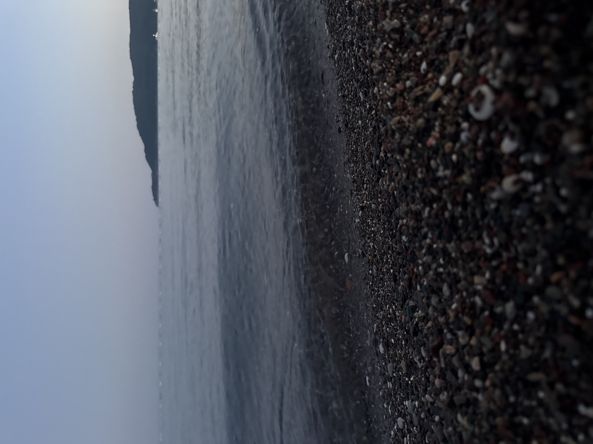 The image depicts a shoreline with a pebble beach in the foreground. The water, appearing calm with small waves lapping at the shore, occupies the midground. In the background, a hazy, tree-covered hill is visible on the horizon, against a pale, twilight sky. The overall impression is one of a serene, natural environment, likely captured at dusk or dawn. The location is identified as Boreti, Montenegro. There are no people, animals, or visible man-made structures in the frame.