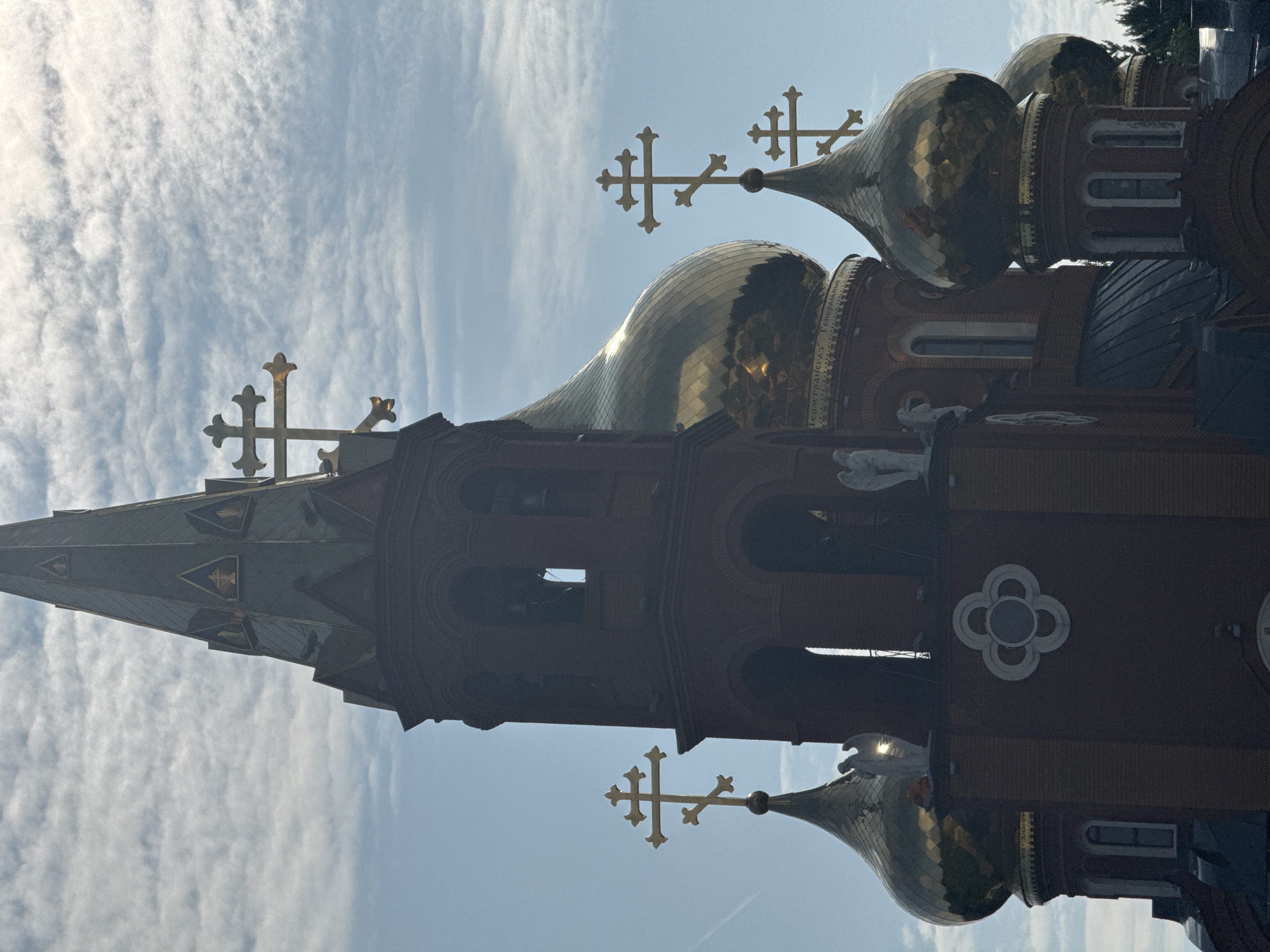 The image shows a close-up view of the spire and domes of the Holy Trinity Cathedral in Chaikovsky, Russia. The cathedral is constructed of red brick and features several golden domes topped with Orthodox crosses. One prominent spire is visible, extending upwards into a sky filled with scattered clouds. The domes are intricately detailed, reflecting the surrounding sky and light. Statues of angels are positioned on ledges near the domes. The overall scene is a detail-oriented shot of the architectural elements of the cathedral against a partly cloudy sky.