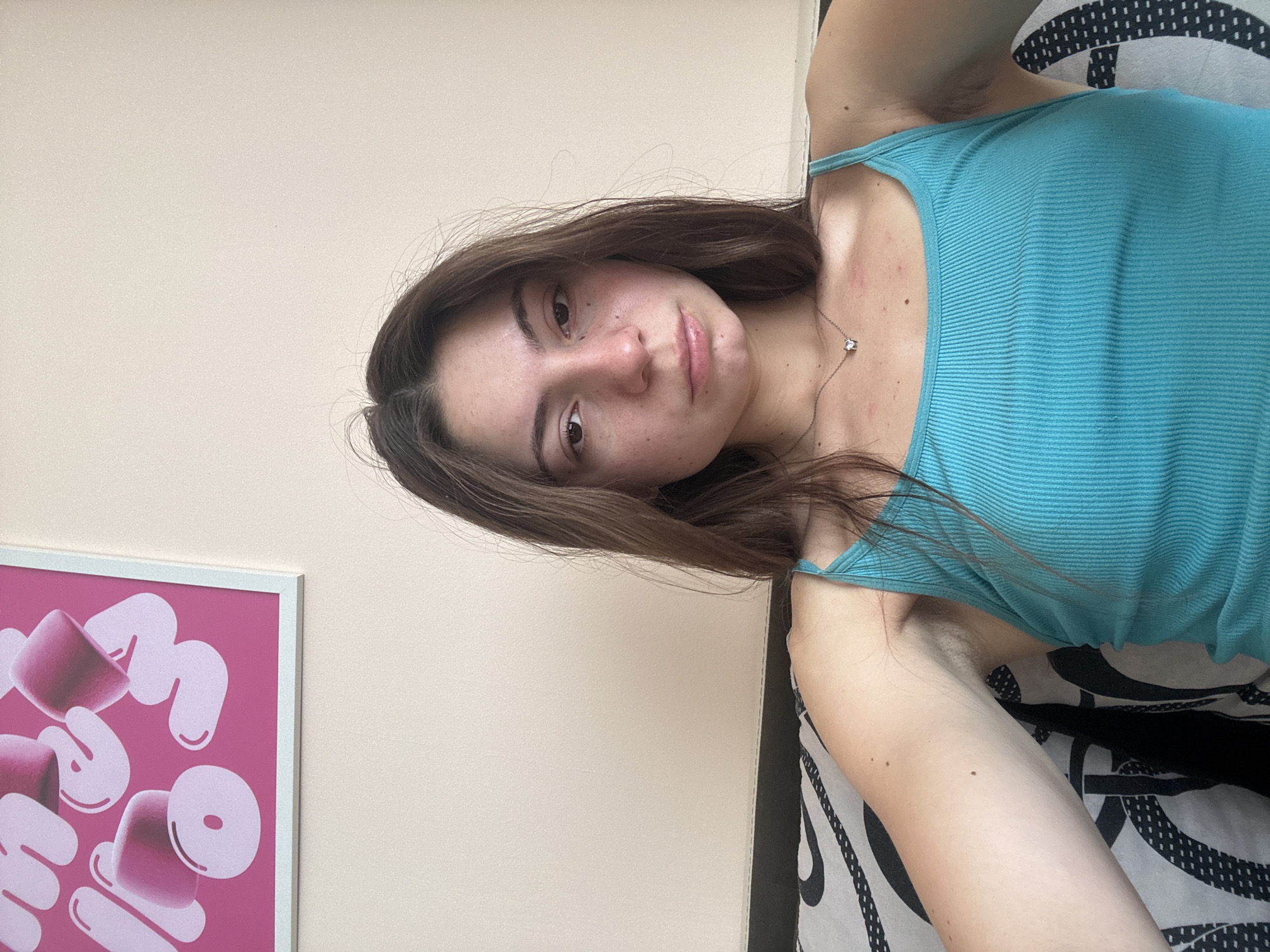 The image shows a young woman with long brown hair, wearing a teal tank top and a silver necklace with a small pendant. She is sitting on a couch with a patterned black and white throw pillow. A framed picture with pink abstract shapes hangs on the wall behind her. The woman is looking directly at the camera with a neutral expression. There are some red marks on her upper chest and shoulder area. The overall lighting suggests it is daytime and the scene appears to be indoors, likely a living room. The city of Varna, Bulgaria is not explicitly depicted or referenced in the image itself.