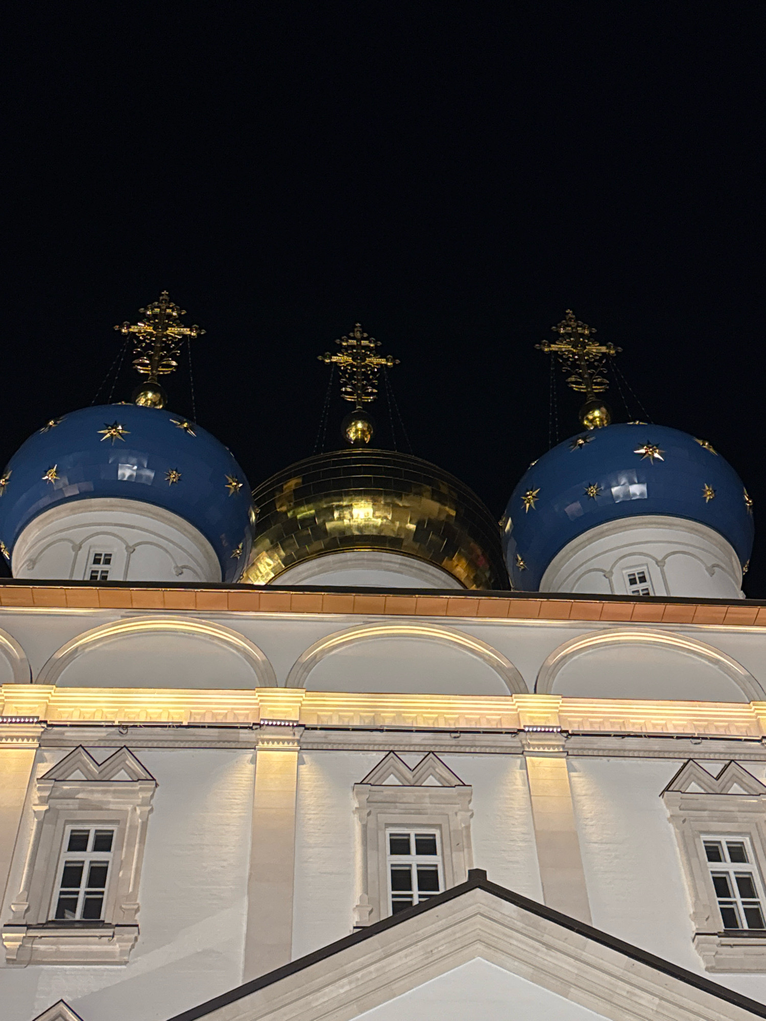 The image shows a nighttime view of the domes of the Ascension Cathedral in Tver, Russia. Three domes are visible, two are bright blue with gold stars, and one is golden. Each dome is topped with a gold cross. The cathedral's white facade is illuminated by warm yellow lights, highlighting architectural details such as arched windows and decorative moldings. The sky above is dark and black, indicating it is nighttime. There are no people visible in the image. The focus is on the exterior architecture of the cathedral, emphasizing its religious and decorative elements.