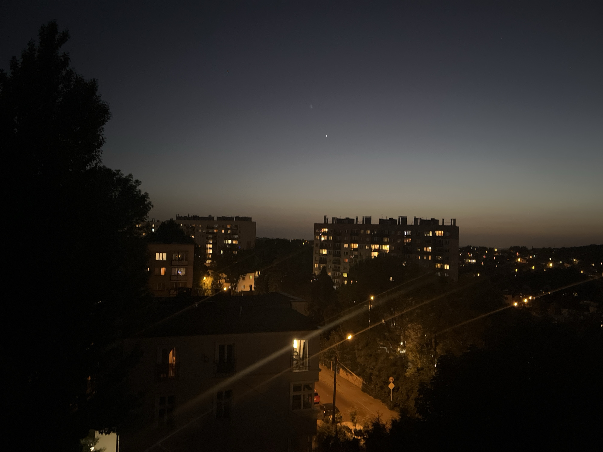 The image shows a cityscape at night. Two large apartment buildings with illuminated windows are visible in the background. A smaller building is in the foreground, also with some illuminated windows. Streetlights line a road that runs through the scene. The sky is dark, with a gradient from a lighter hue near the horizon to a darker color above. Trees are silhouetted on the left and right sides of the image. The location appears to be a residential area of a city.