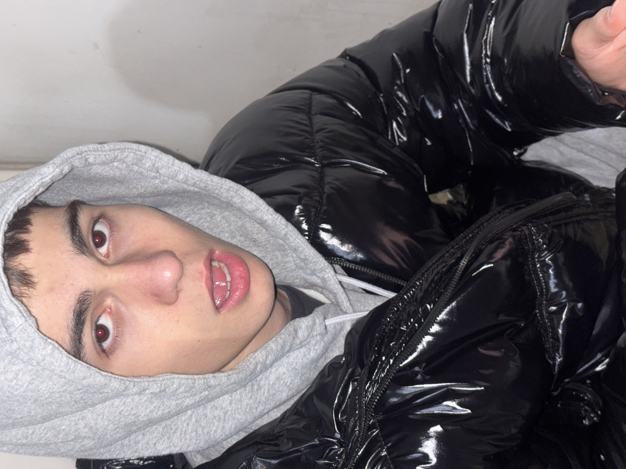 A young man is lying down, wearing a grey hoodie and a shiny black puffer jacket. The location is unknown and cannot be determined from this image.