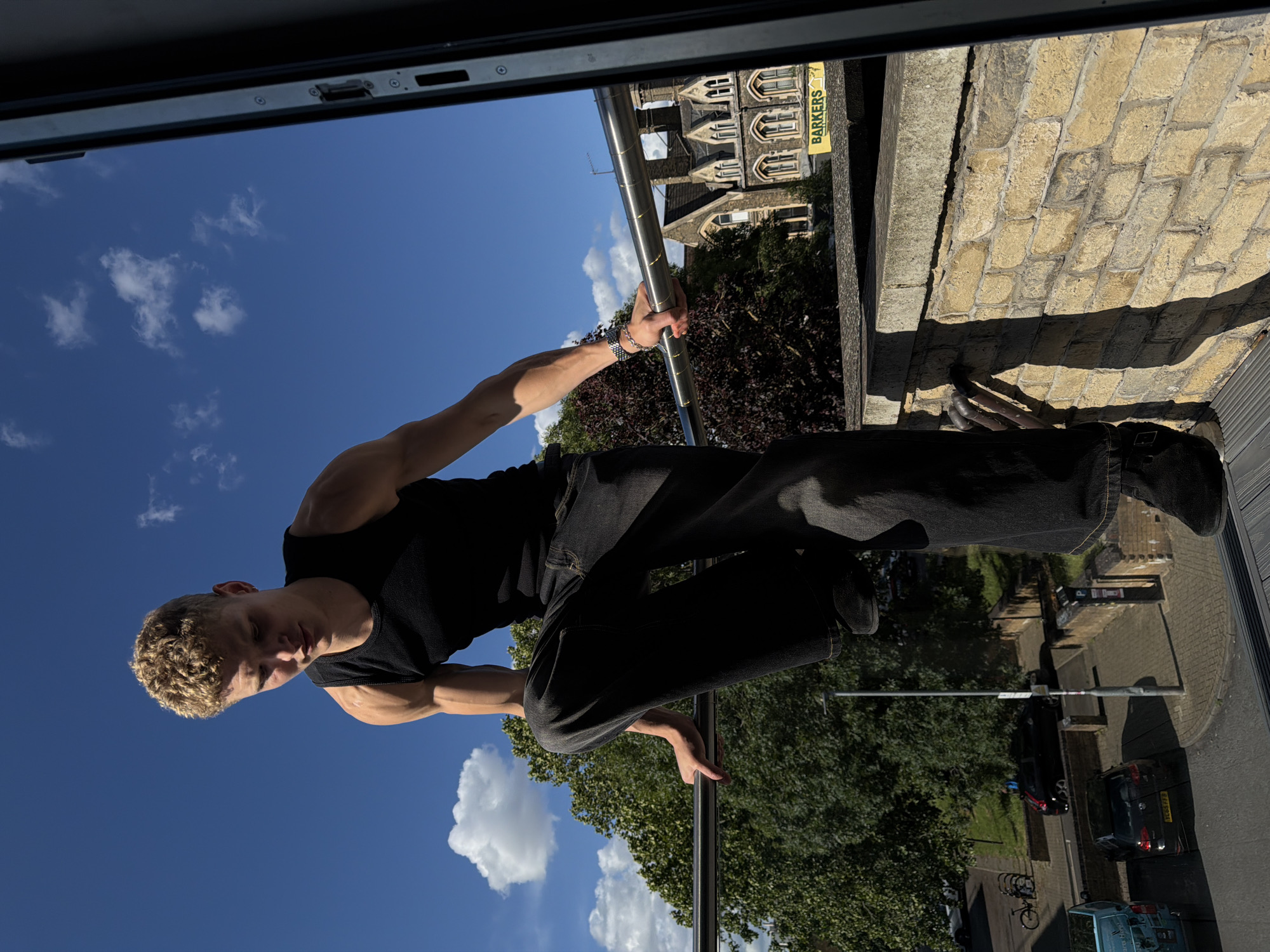 A young man with short, curly blonde hair and fair skin, wearing a black tank top and dark wash wide-leg jeans, is perched on a metal railing outdoors. He is looking down, his arms resting on the railing. He has a silver watch on his left wrist. The background features a bright blue sky with scattered white clouds, lush green trees, and a building with multiple windows and a prominent yellow sign that reads "BARKERS". The man appears to be on a balcony or rooftop in an urban environment. Below him, a street is visible with parked cars and a bicycle rack. The overall lighting suggests it is daytime, with strong sunlight casting shadows.