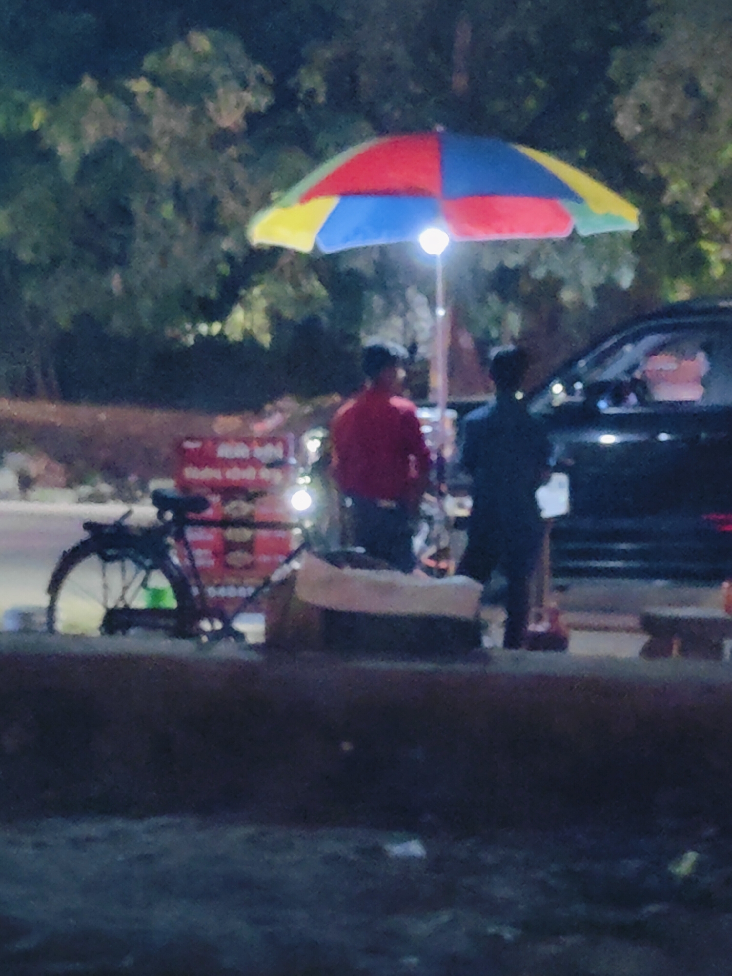 The image depicts a nighttime scene in Gurgaon, India, likely near a roadside market or food stall. A colorful umbrella, with sections of red, yellow, and blue, is set up, supported by a pole that also has a bright light attached. Underneath the umbrella, two individuals are visible. One person, wearing a red shirt, is standing near a bicycle with a basket and what appears to be a food cart or stall with red signage. The other person, wearing a dark blue shirt, is standing next to a black car, seemingly interacting with someone inside the vehicle. The background is filled with dark foliage, suggesting trees or bushes lining the area. The foreground shows a dark, out-of-focus surface that could be a road or pathway. The lighting is dim, with the exception of the umbrella light and the car's headlights, creating a somewhat grainy and indistinct image.