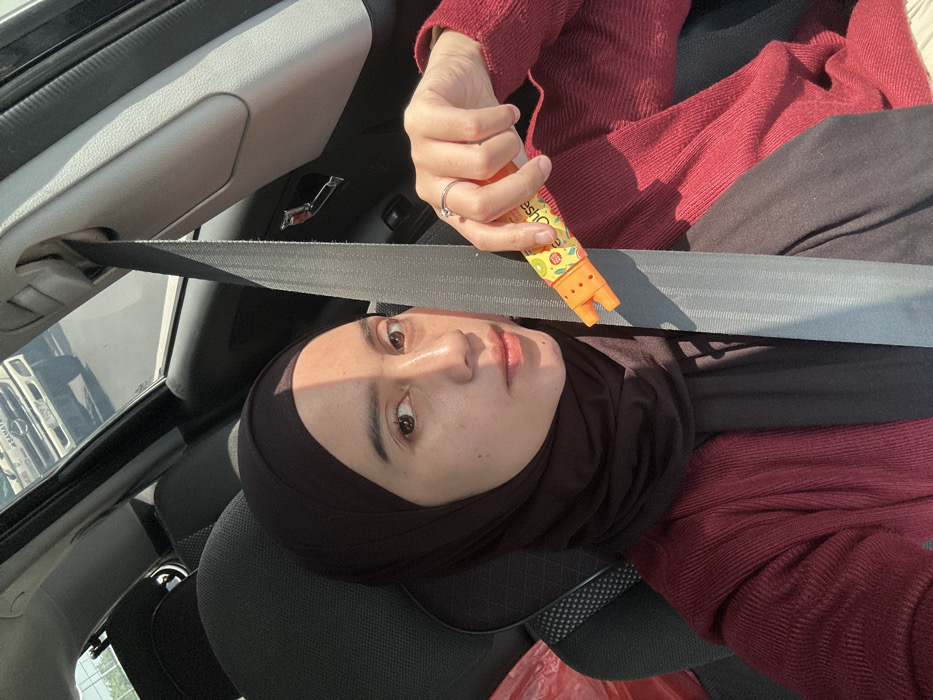 The image depicts a single individual, likely female, seated in the front passenger seat of a vehicle. She is wearing a dark brown hijab and a dark reddish-brown ribbed cardigan over a dark inner garment. A grey seatbelt is fastened diagonally across her chest. Her right hand, adorned with a silver ring on the ring finger, holds an orange and yellow dispenser-like object, possibly a candy dispenser, featuring fruit graphics and partial text "Juice." Her face is partially illuminated by direct sunlight from the right, casting a shadow across her left side. In the background, visible through the car window, is a white commercial truck, possibly with "SAMATRA" (or similar partially visible text) on its grille and a "B" prefix on its license plate, suggesting an Indonesian registration. The vehicle appears to be on a street or road during daylight hours. This scene is situated in the city of Entalsewu, Indonesia.