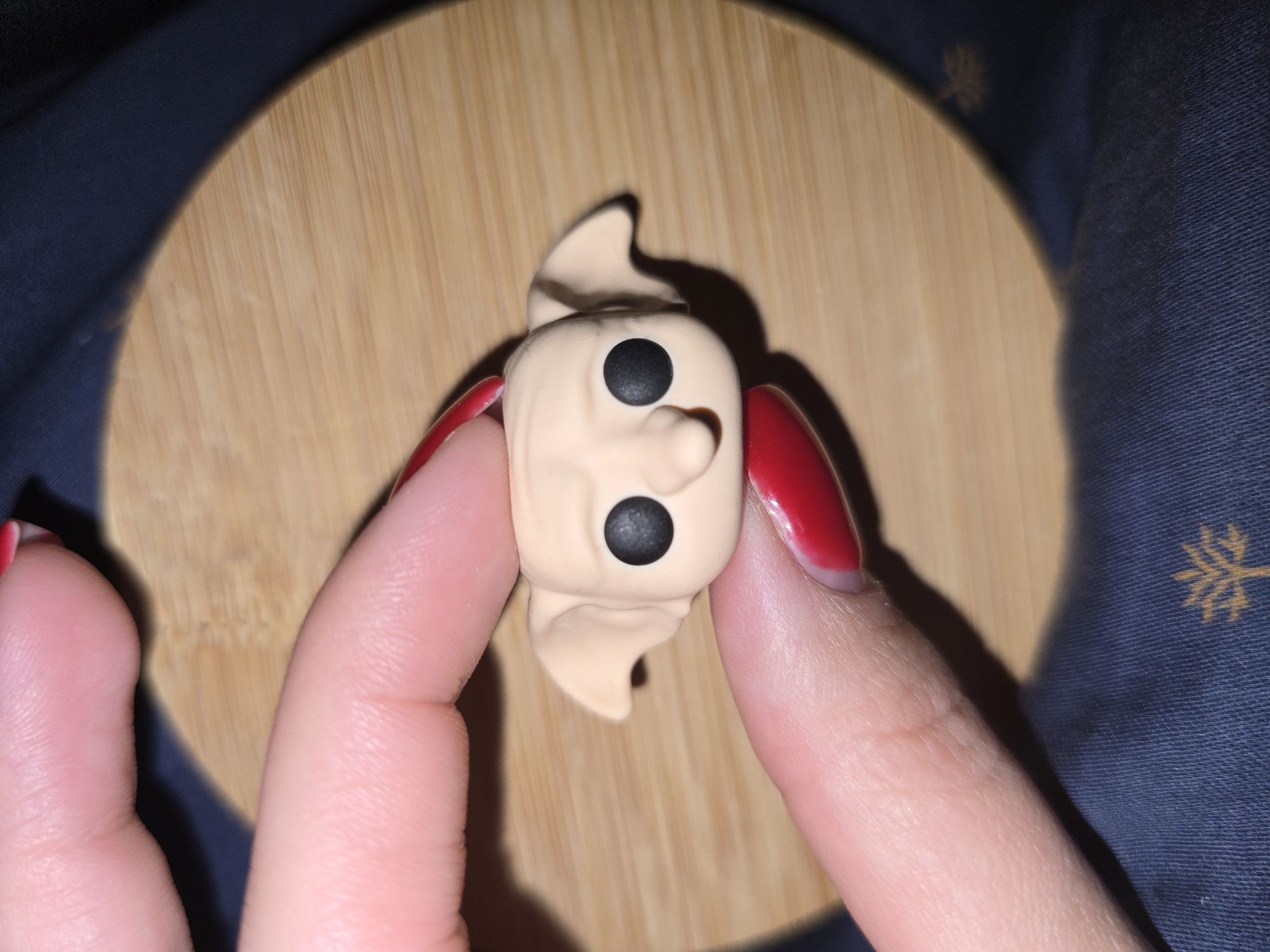 A pair of human fingers, with red nail polish, hold a small, tan-colored doll with large black eyes and prominent ears. The doll appears to be a Funko Pop figure, possibly representing the character Dobby from Harry Potter. The doll is positioned over a round, light-brown wooden surface, which is likely a coaster or placemat. In the background, a dark blue fabric with a faint yellow pattern is visible. The lighting in the image creates shadows and highlights on the doll and fingers. The overall setting suggests a close-up indoor shot. The image does not contain any information related to the city of Petropavlivska Borshchahivka, Ukraine.