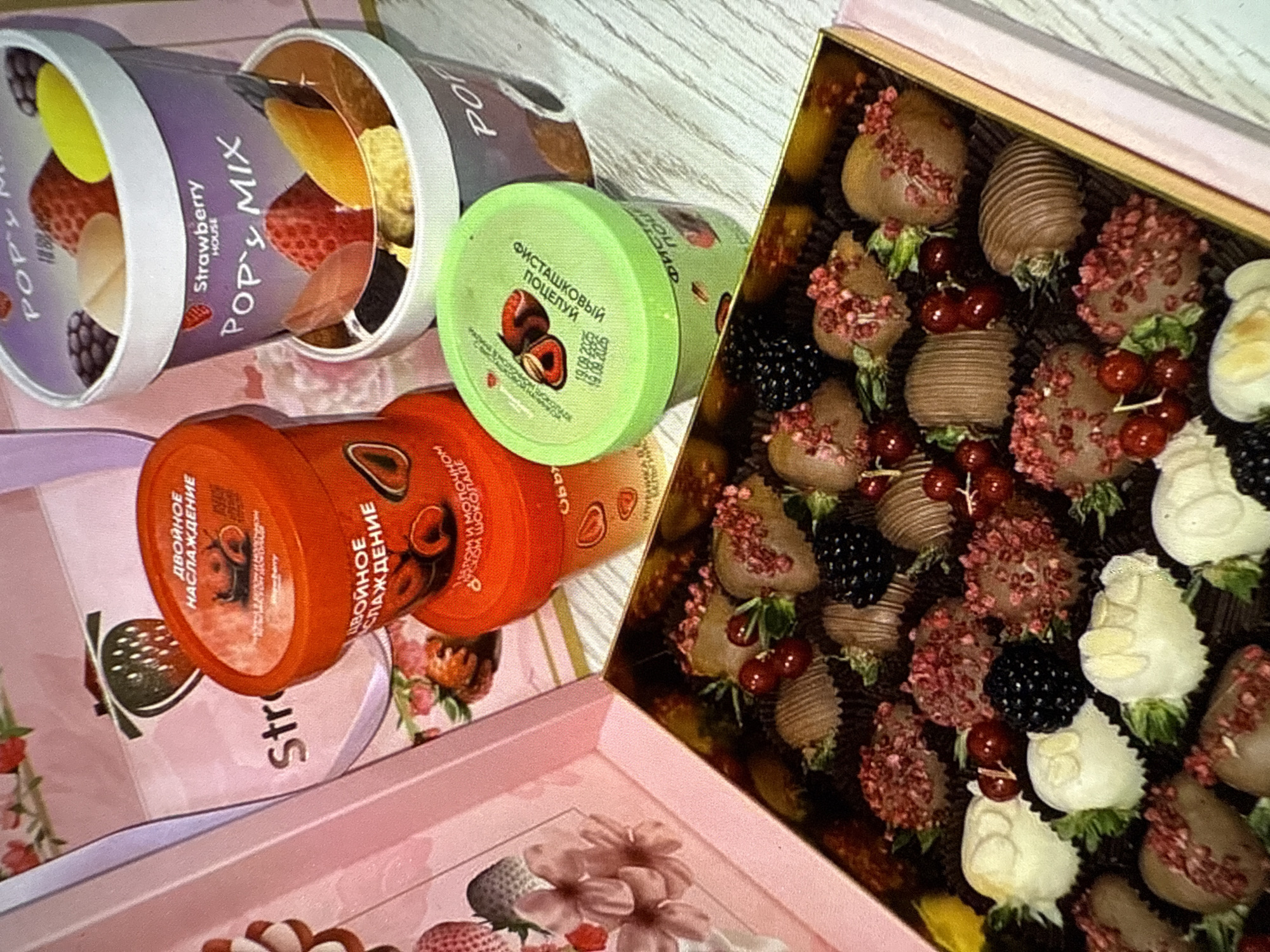 A top-down, medium shot captures a meticulously arranged box of assorted chocolate-covered strawberries and other confectionery items. The box itself is a pale pink, rectangular container with a gold interior, showcasing an array of treats. The chocolates are varied in design, with some being heart-shaped and covered in milk chocolate with red sprinkles, while others are coated in white chocolate with intricate detailing. Blackberries and red currants are interspersed amongst the chocolates, adding pops of color and texture.
In the background, several cylindrical containers are visible. Two are purple with white lids, featuring colorful graphics of strawberries and other fruits, and text that reads "POP's MIX" and "Strawberry HOUSE." To the left of these, a bright orange cylindrical container is present, with similar strawberry graphics and Cyrillic text, possibly indicating "DOUBLE DELIGHT." A green cylindrical container with white lid, also with fruit imagery and Cyrillic text, is situated in the foreground, to the right of the orange one.
The overall presentation suggests a gift or a special treat, with the confectionery items arranged with care. The lighting is bright and even, highlighting the details of the food items and packaging. The environment appears to be a flat, textured surface, possibly a table, with a subtle pattern. No people or any specific location details are discernible in the image.