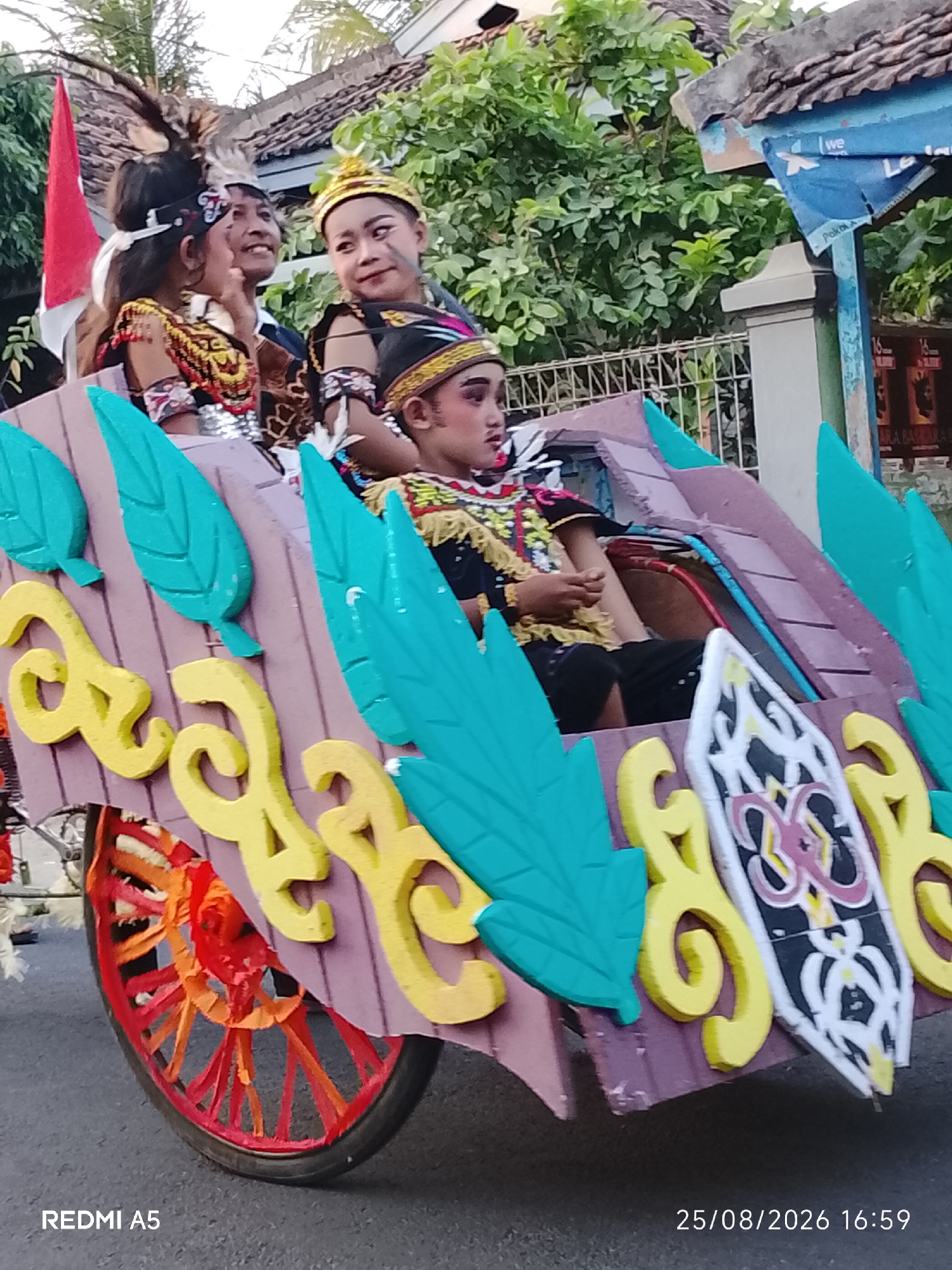 This is a medium shot, outdoor, eye-level, full-color photograph of a parade float with several individuals dressed in traditional attire. The float is constructed of wood and painted in shades of purple, yellow, and teal, with decorative elements resembling leaves and ornate carvings. A red-wheeled cart is visible at the front of the float.
Four individuals are visible on the float. On the left, a young female wears a headdress with feathers and a colorful, beaded garment. Next to her, a male smiles warmly and looks towards the left. To his right, another young female with a golden crown and striking eye makeup is seated. In the front of the float, a young male is seated, also in traditional attire, looking forward.
In the background, lush green foliage and trees suggest a tropical environment. A red and white flag is visible on the left side of the float, possibly indicating a national or regional affiliation. A blue sign with white lettering is partially visible on the right side, and what appears to be a building or structure with a tiled roof is also present. The ground is paved with asphalt. The time stamp in the lower right corner indicates the date and time as 25/08/2026 16:59, and the device used to capture the image is identified as REDMI A5 in the lower left corner. The location is identified as Semanding, Indonesia.