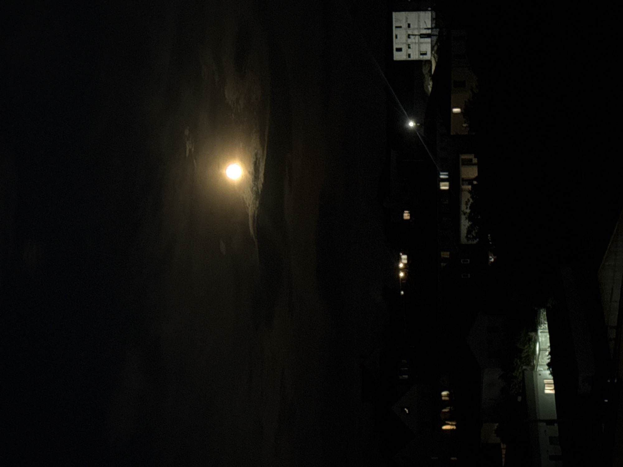 The image depicts a nighttime scene of a residential area in Балаклавский округ, Ukraine. A bright, full moon is visible in the upper portion of the frame, partially obscured by scattered clouds. Below the moon, the dark silhouettes of various buildings and houses are discernible. Some windows in these structures are illuminated, suggesting occupancy. A few streetlights are also visible, casting a faint glow on the surroundings. The overall impression is a quiet, residential neighborhood under the moonlight.