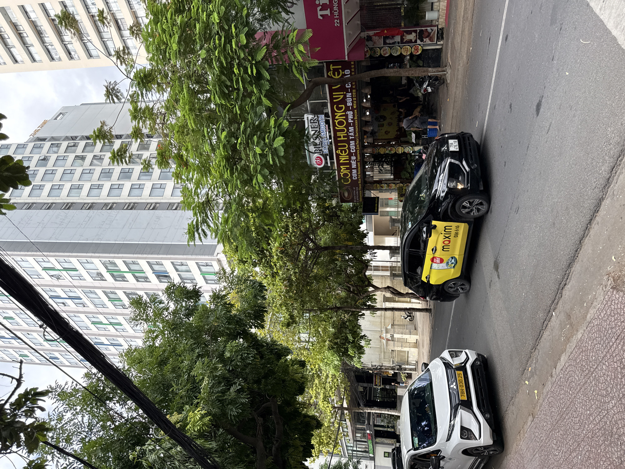 The image depicts a street scene in Nha Trang, Vietnam, characterized by a multi-lane road bordered by buildings and lush trees. In the foreground, a white Mitsubishi Xpander taxi with the license plate "78H-030.25" is parked on the right side of the road, facing left. To its right, a black and yellow Maxim taxi, also identified as a Mitsubishi Xpander with the license plate "79A 476.18", is moving forward. The Maxim taxi features prominent branding, including the "maxim" logo and advertisements for ride-hailing services.
The environment includes a sidewalk with a patterned surface and a street with a white dividing line. Several buildings line the street, including a tall, modern residential or hotel building with a repetitive window pattern and a smaller building with commercial establishments. One of these businesses is "CƠM NIÊU HƯƠNG VỊ VIỆT", a Vietnamese rice pot restaurant, indicated by a large sign with text in Vietnamese and other languages. Another establishment displays the "PILSNER EXTRA SMOOTH" logo. Trees with dense green foliage are scattered throughout the scene, providing shade and contributing to the urban landscape. The sky is overcast, suggesting a cloudy day. A few individuals are visible, including one person walking near the buildings and another standing outside the restaurant. A motorcycle is also parked on the sidewalk. Power lines are visible overhead, adding to the urban infrastructure.