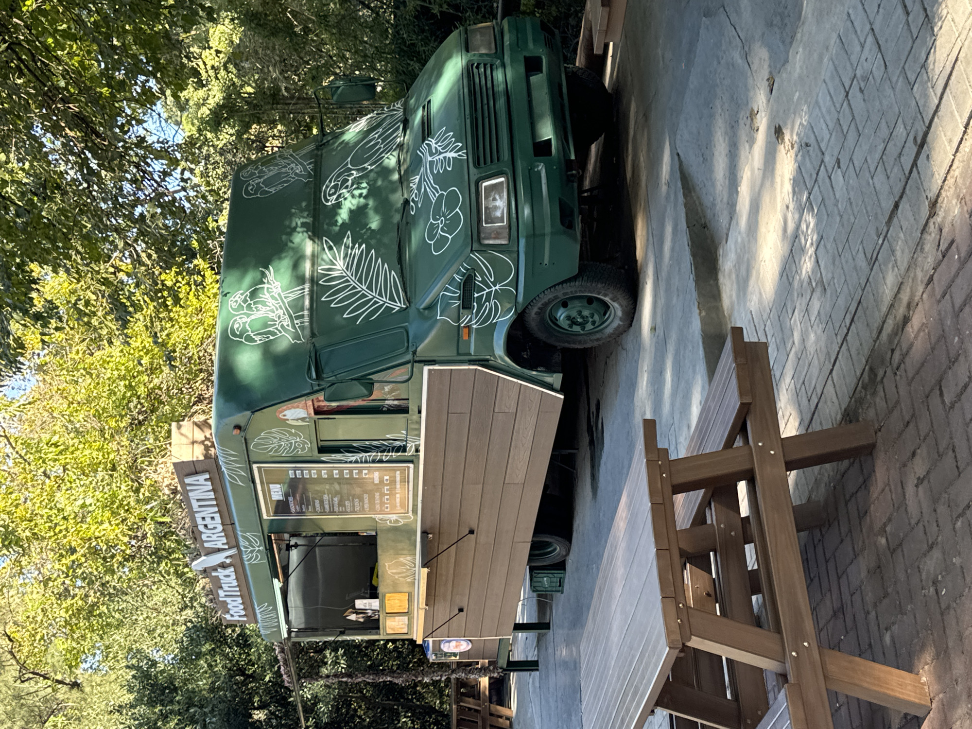 A green food truck is parked outdoors on a paved area, adjacent to wooden tables and benches. The food truck is decorated with white drawings of plants and birds and features a sign that reads "Food Truck Argentina." The menu displayed includes items like sandwiches, salads, juices, and desserts. The truck is likely serving Argentinian cuisine. The scene appears to be set in a park or outdoor area in Buenos Aires, Argentina, based on the signage on the truck. There are no people visible in the immediate frame. The surroundings consist of trees and greenery. The ground is a mix of concrete and brick paving.