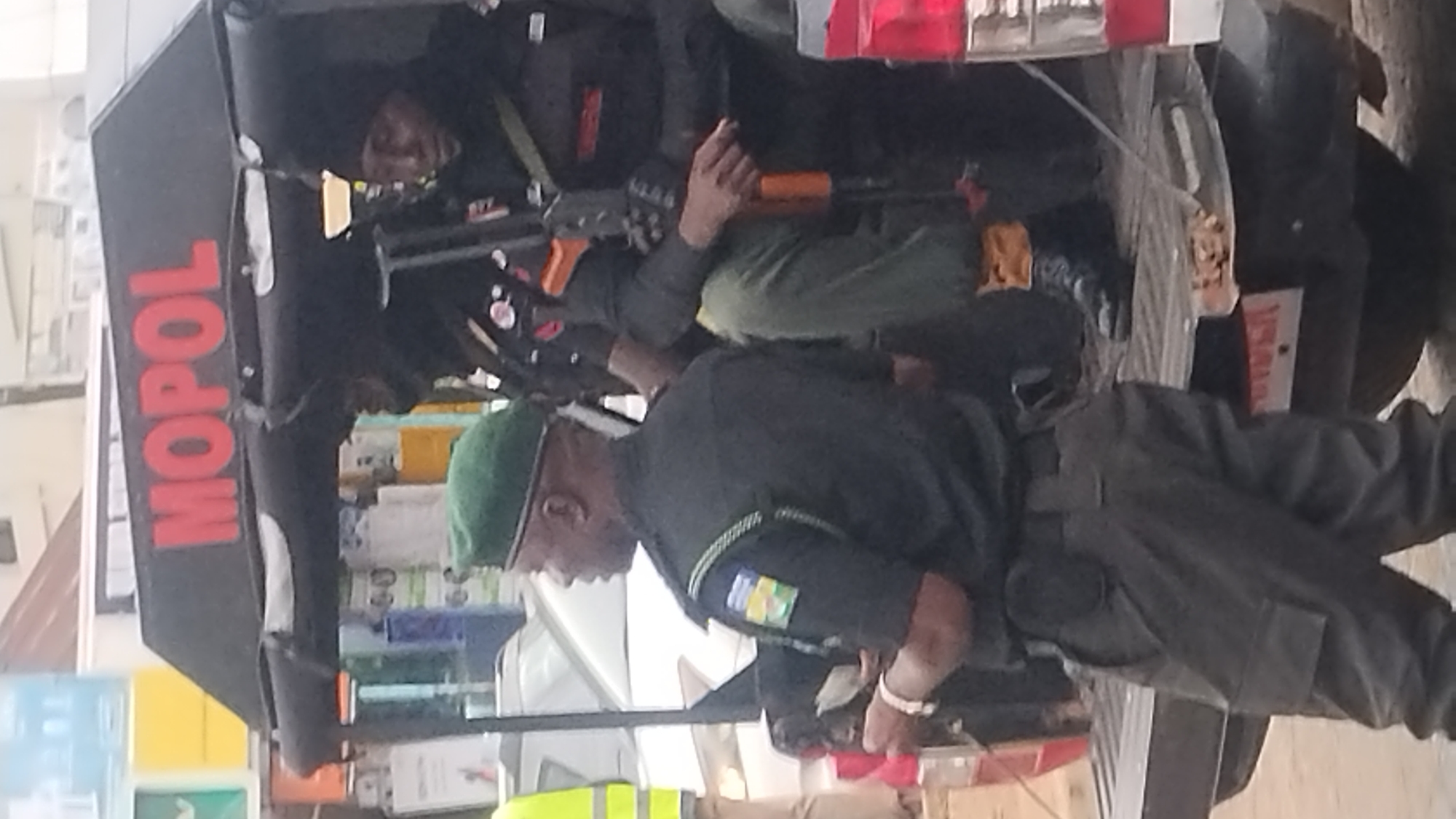 The image shows several individuals in what appears to be a busy outdoor setting in Jos, Nigeria. The primary subjects are individuals dressed in what looks like law enforcement or security uniforms, some of whom are holding firearms. The vehicles in the background have "MOPOL" written on them in red letters, which is an acronym for Mobile Police.
The scene depicts a group of individuals, likely police officers, engaged in some form of operational activity. One person is holding a rifle with an orange foregrip. Another individual, wearing a green beret, is seen in the foreground looking down. The overall impression is one of readiness or ongoing duty.
The setting appears to be an urban environment, with elements of what could be shops or stalls in the background, indicated by shelves stocked with various items. The lighting suggests it is daytime. There are no clear indicators of specific weather conditions. The presence of firearms and uniformed personnel suggests a security-related event or patrol.
The text "MOPOL" is clearly visible on the side of a vehicle. There might be other text on signs or clothing, but it is not easily decipherable due to the image quality and orientation. The image is oriented vertically, with the bottom of the scene appearing at the top.