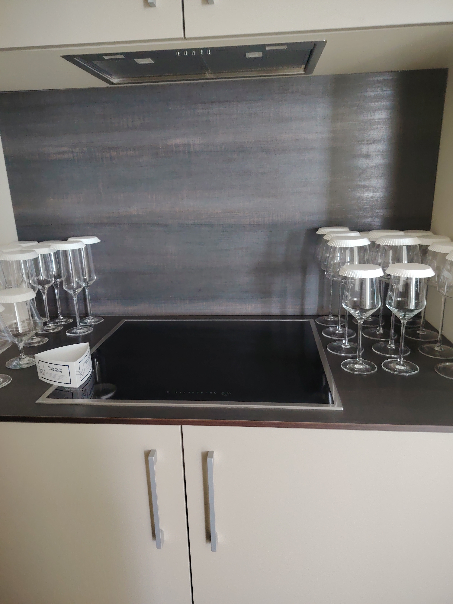 The image shows a modern kitchen counter with a black induction stovetop. To the left of the stovetop, several wine glasses with white covers are neatly arranged. A small white triangular box with text on it is placed in front of the glasses. The background features a dark, textured backsplash. Below the counter are two light-colored cabinet doors with silver handles. The scene appears to be an indoor setting, likely a kitchen, but there is no indication of the city of Konigil, Uzbekistan, or any people.