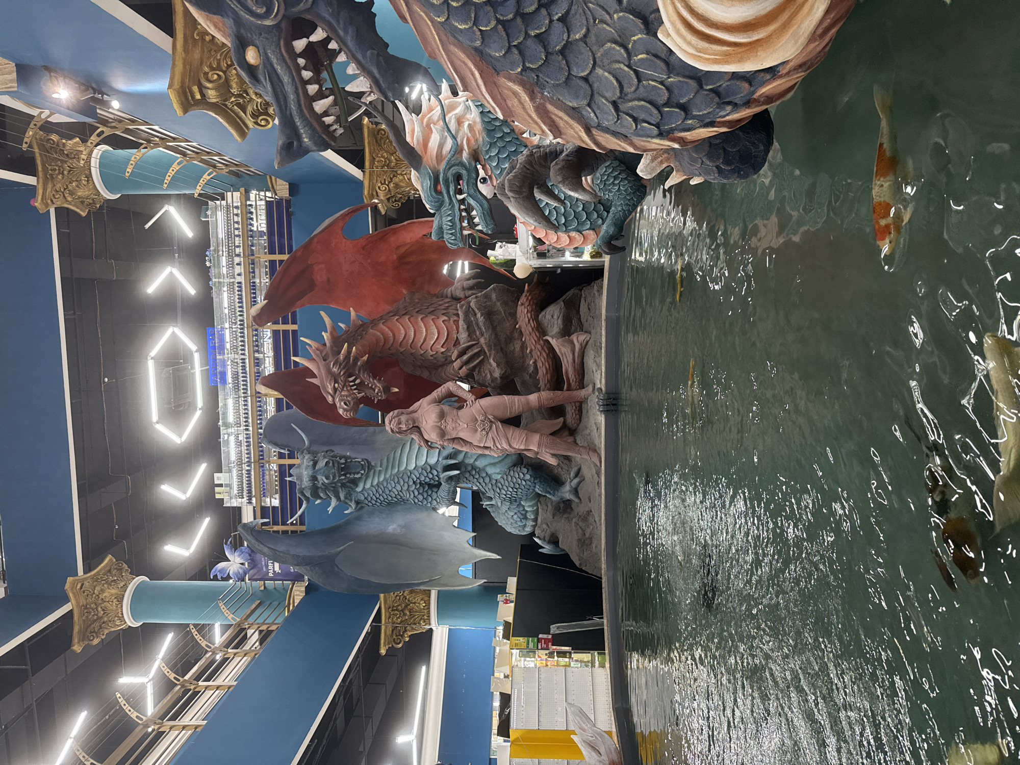 The image shows a multi-level art installation featuring dragons and a human figure positioned above a pool of water with fish. The dragons are sculpted in various colors, including blue, red, and green. The pool reflects light and contains several koi fish. In the background, overhead lighting and shelving units indicate the installation is located within a retail store. Based on information not visible in the image, this location is believed to be in Nakhodka, Russia.