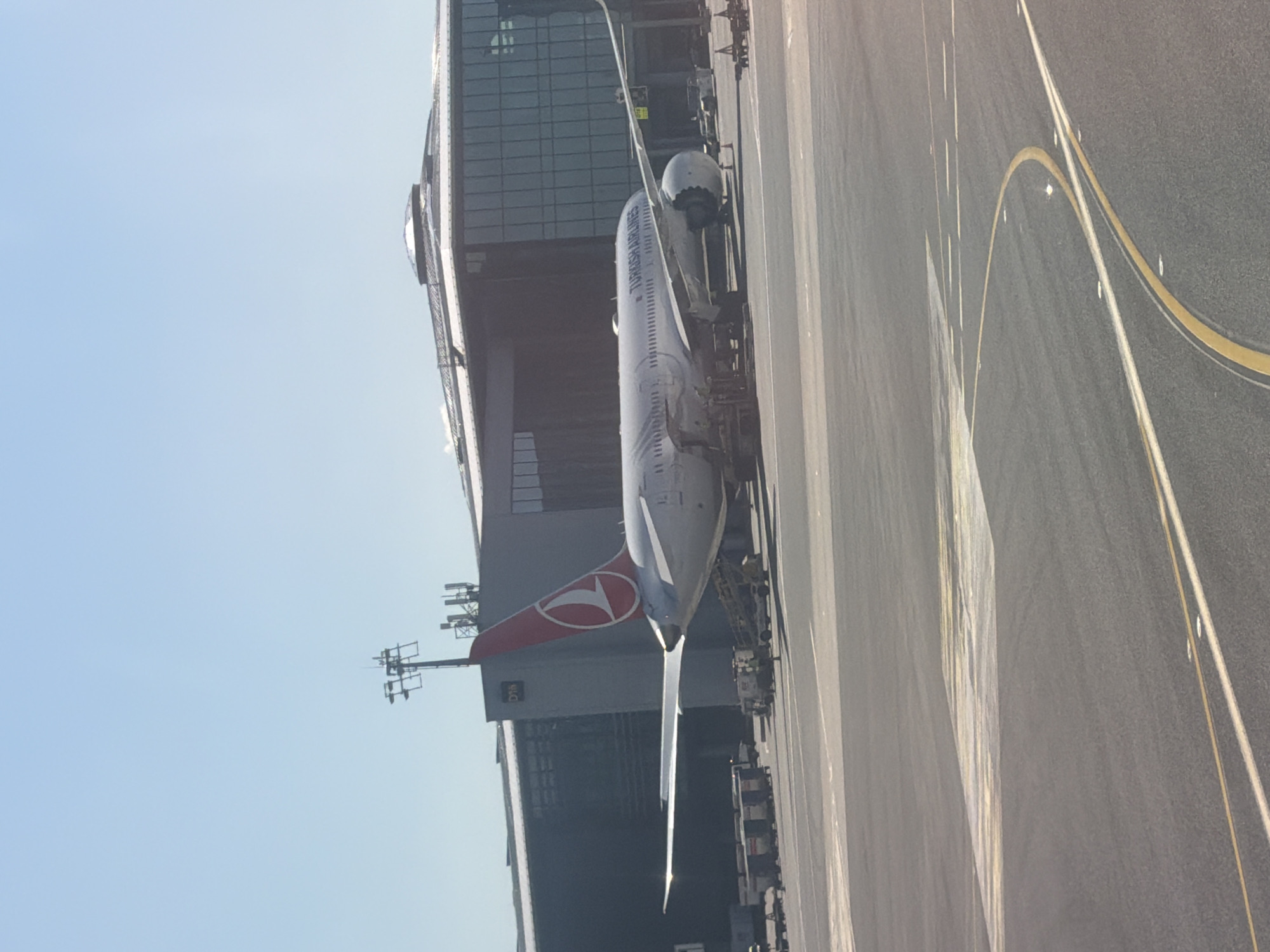 A large Turkish Airlines passenger jet is seen parked at an airport gate under a clear, bright blue sky. The image is taken from a slightly elevated perspective, looking down at the aircraft and the tarmac. The airplane's white fuselage clearly displays the words "TURKISH AIRLINES," and its red tail fin features the airline's distinctive white stylized bird or flower logo.
The aircraft is positioned next to a modern airport terminal building characterized by its extensive glass facade. Various pieces of ground support equipment are visible around the plane, suggesting it is either being prepared for a flight or has recently arrived. The tarmac in the foreground and beneath the aircraft is marked with yellow and white lines, typical of an airport taxiway or apron area. To the left of the terminal building, in the distance, are metal structures resembling communication towers or antenna arrays. The scene depicts a sunny day at an airport, likely during morning or afternoon hours, given the bright illumination.